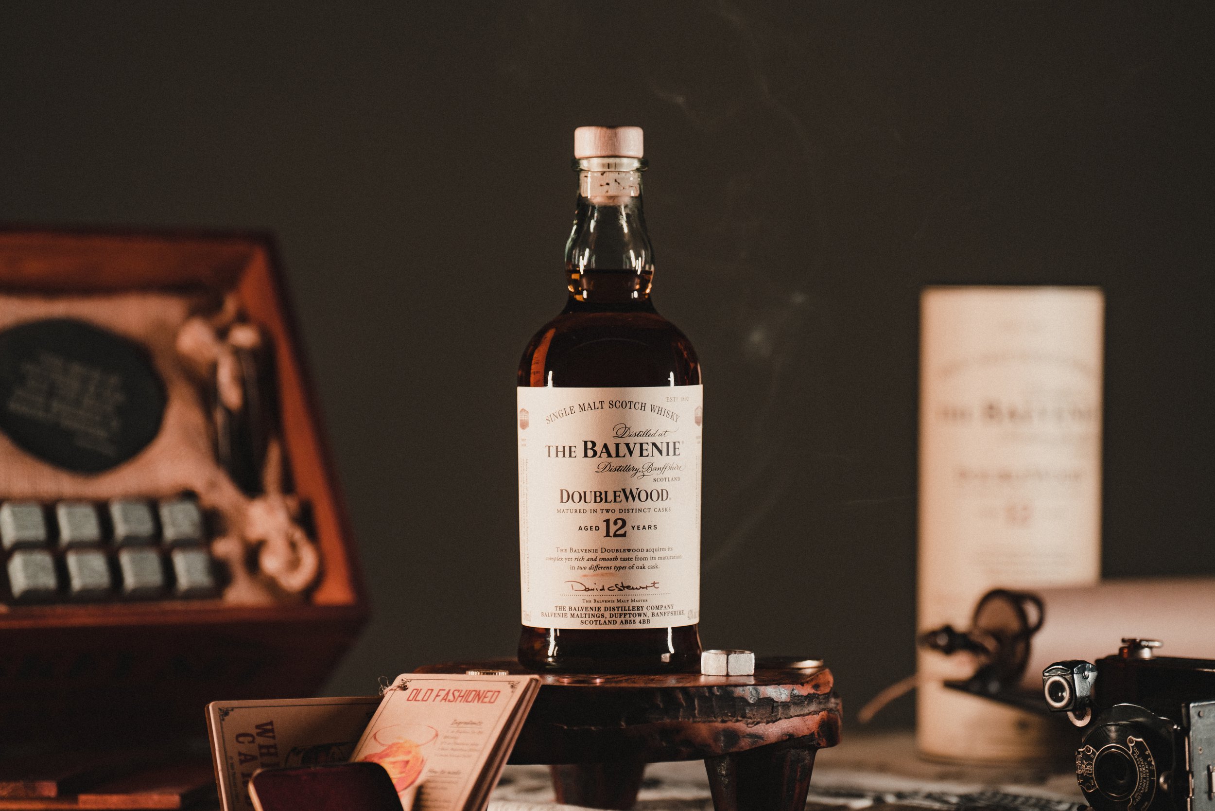 A bottle of Balvenie Double Wood 12-year-old single malt Scotch whisky on a small wooden table, surrounded by vintage items including an old camera, a box with a leather strap, and a menu or pamphlet.