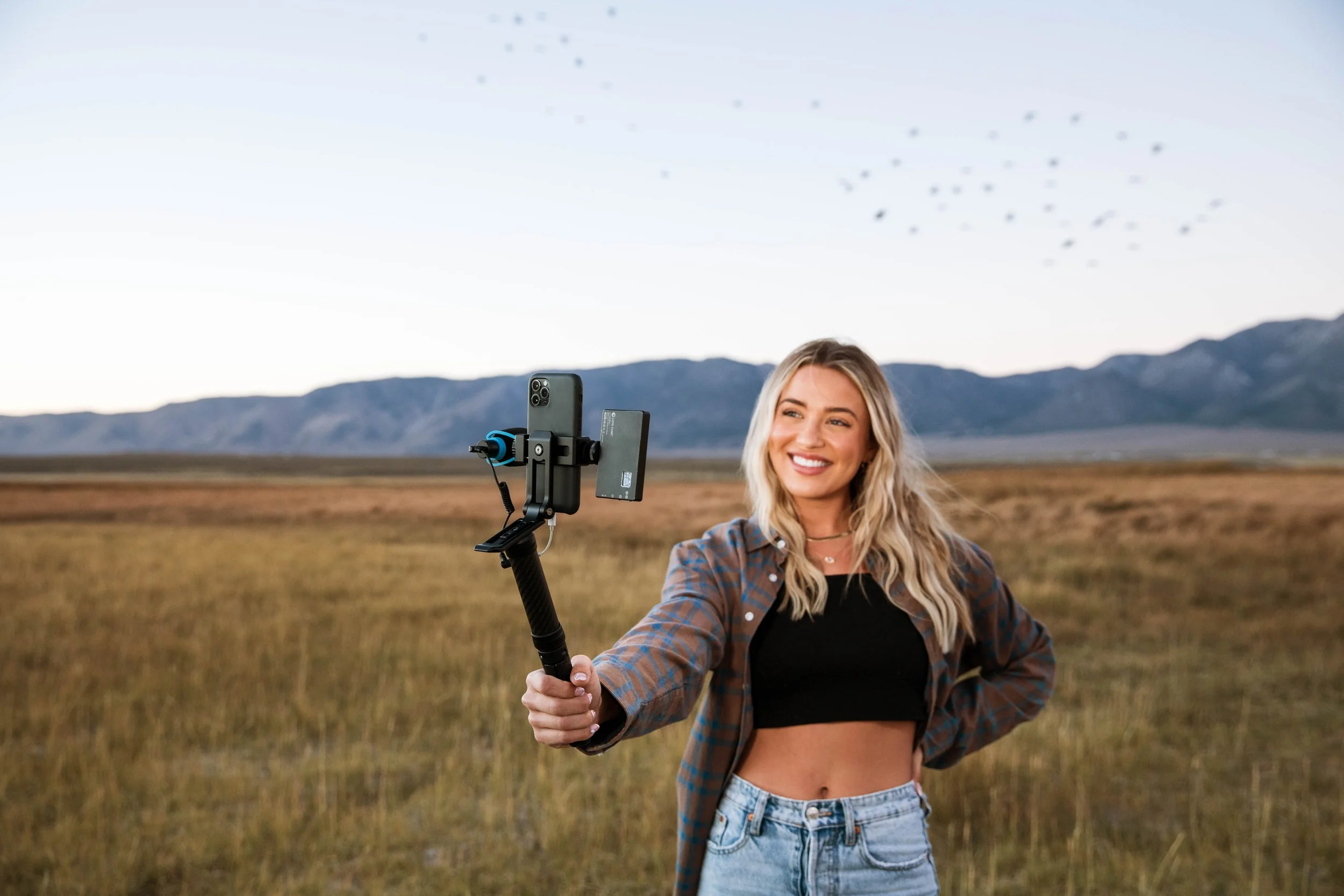 A young woman with blonde hair takes a selfie with a smartphone on a selfie stick in an open field with mountains in the background and birds flying in the sky.