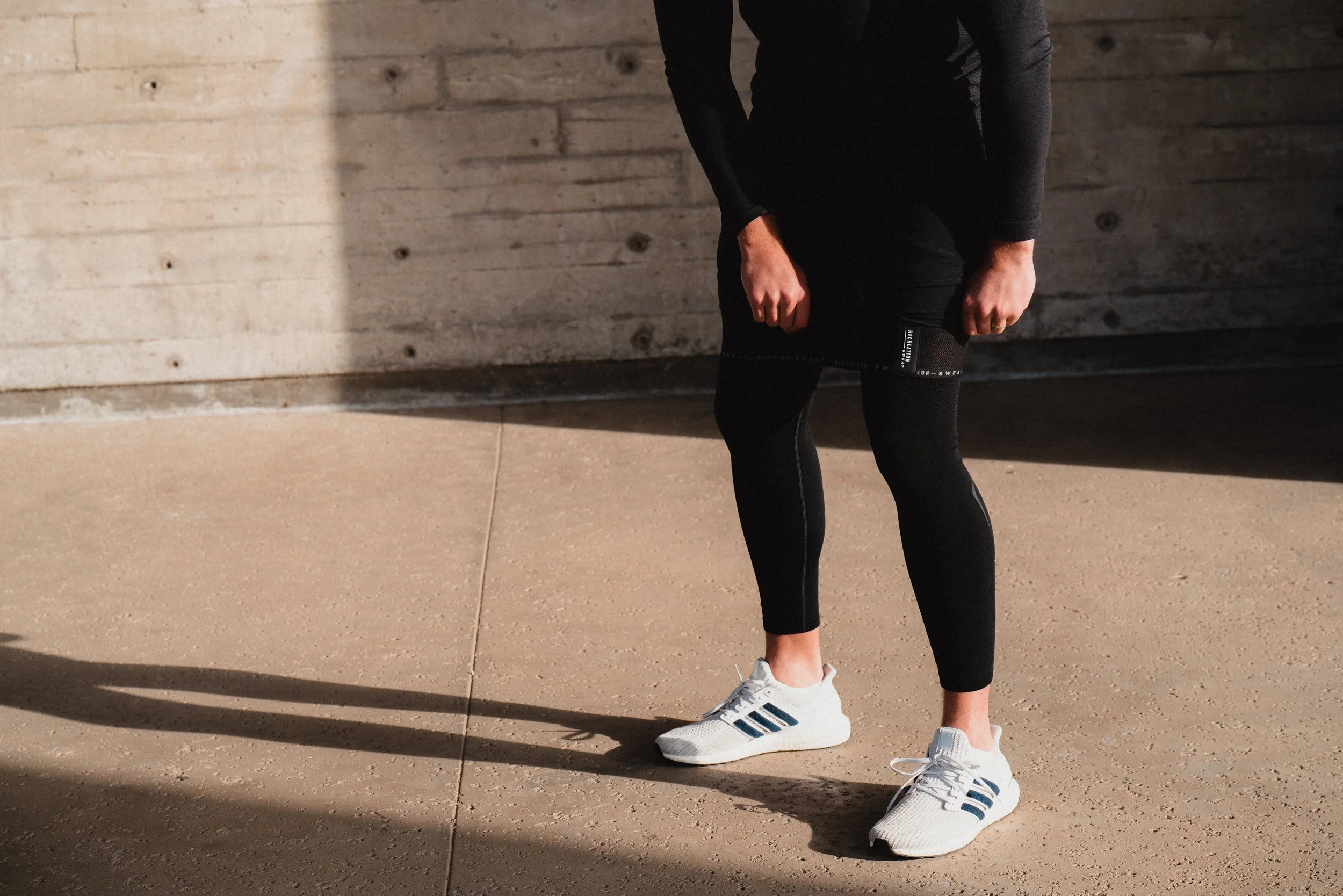 A person dressed in black athletic wear and white sneakers standing on a concrete surface, casting a shadow.