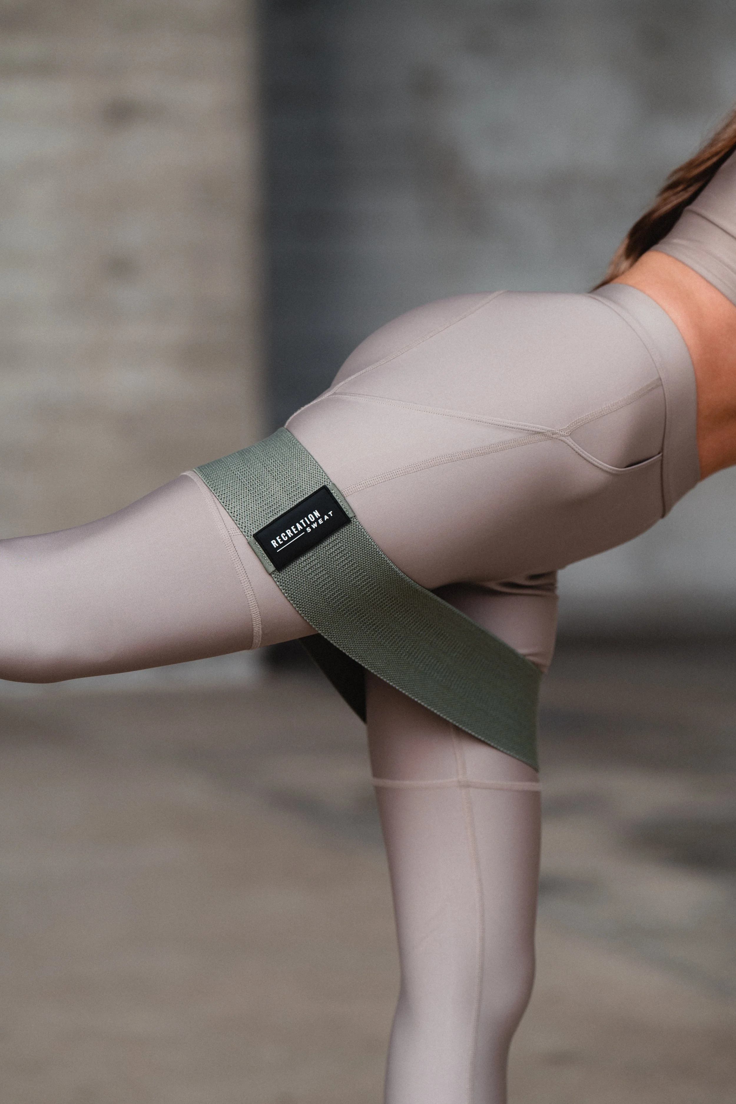 A woman wearing beige activewear with a green resistance band around her thighs, performing a workout move.