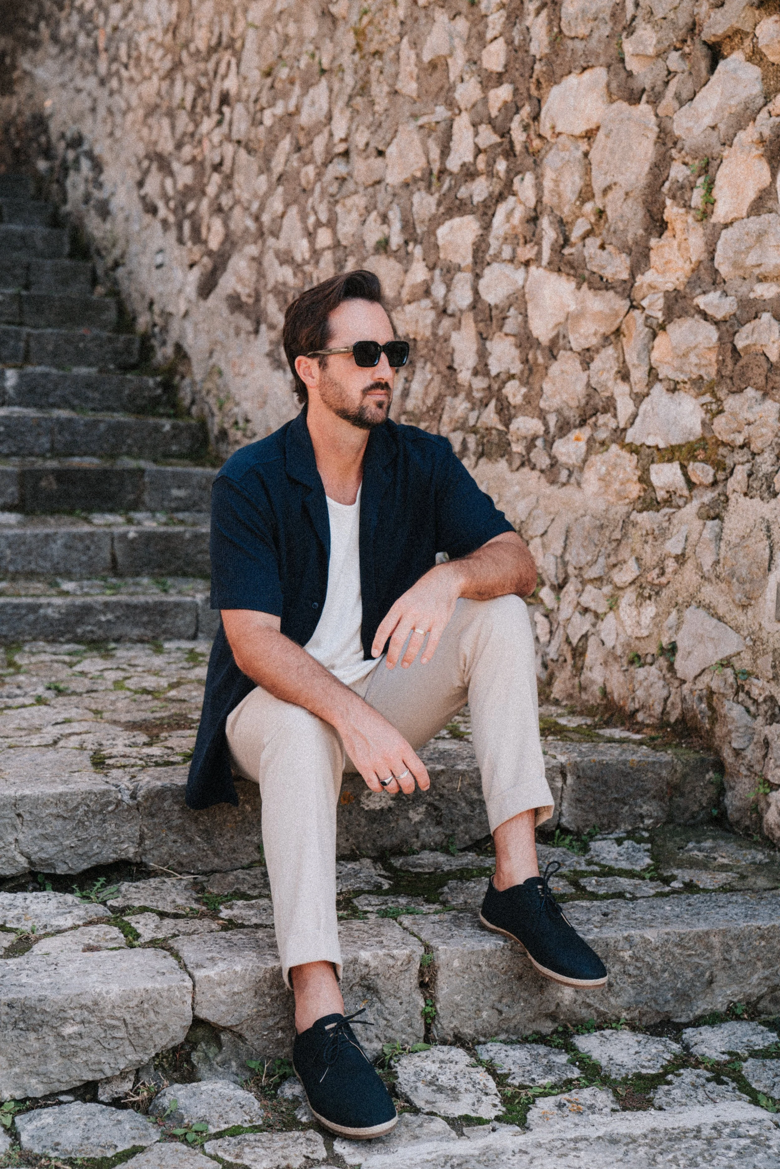 Man sitting on stone steps near a stone wall, wearing sunglasses, a navy blue shirt, beige pants, and black shoes.