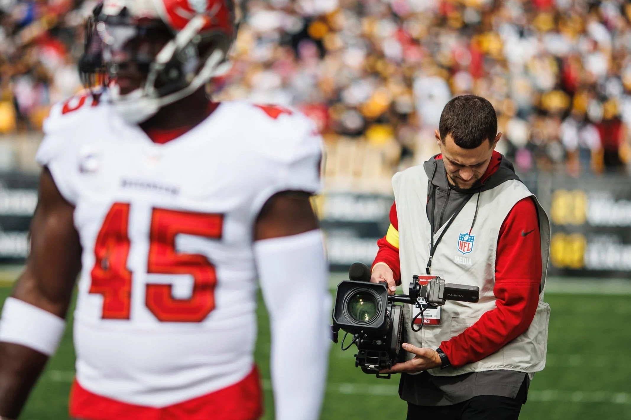 A football player in a white and red uniform with the number 45 on his jersey on the field, with a cameraman filming him nearby during a game.