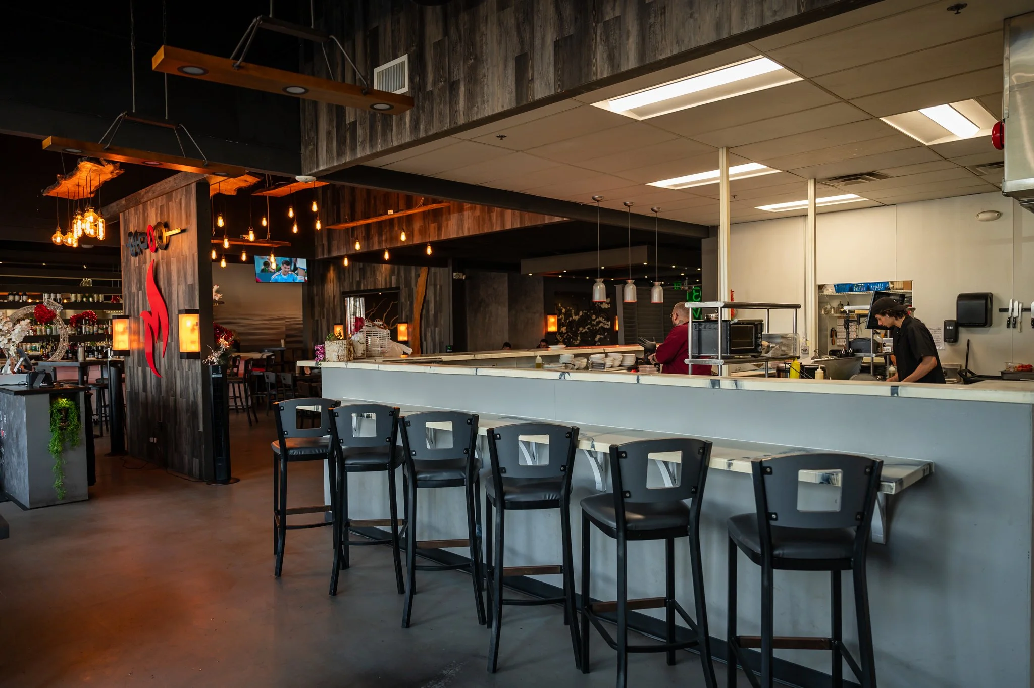 Inside a modern restaurant with a bar area, black chairs, and a chef preparing food in the open kitchen.