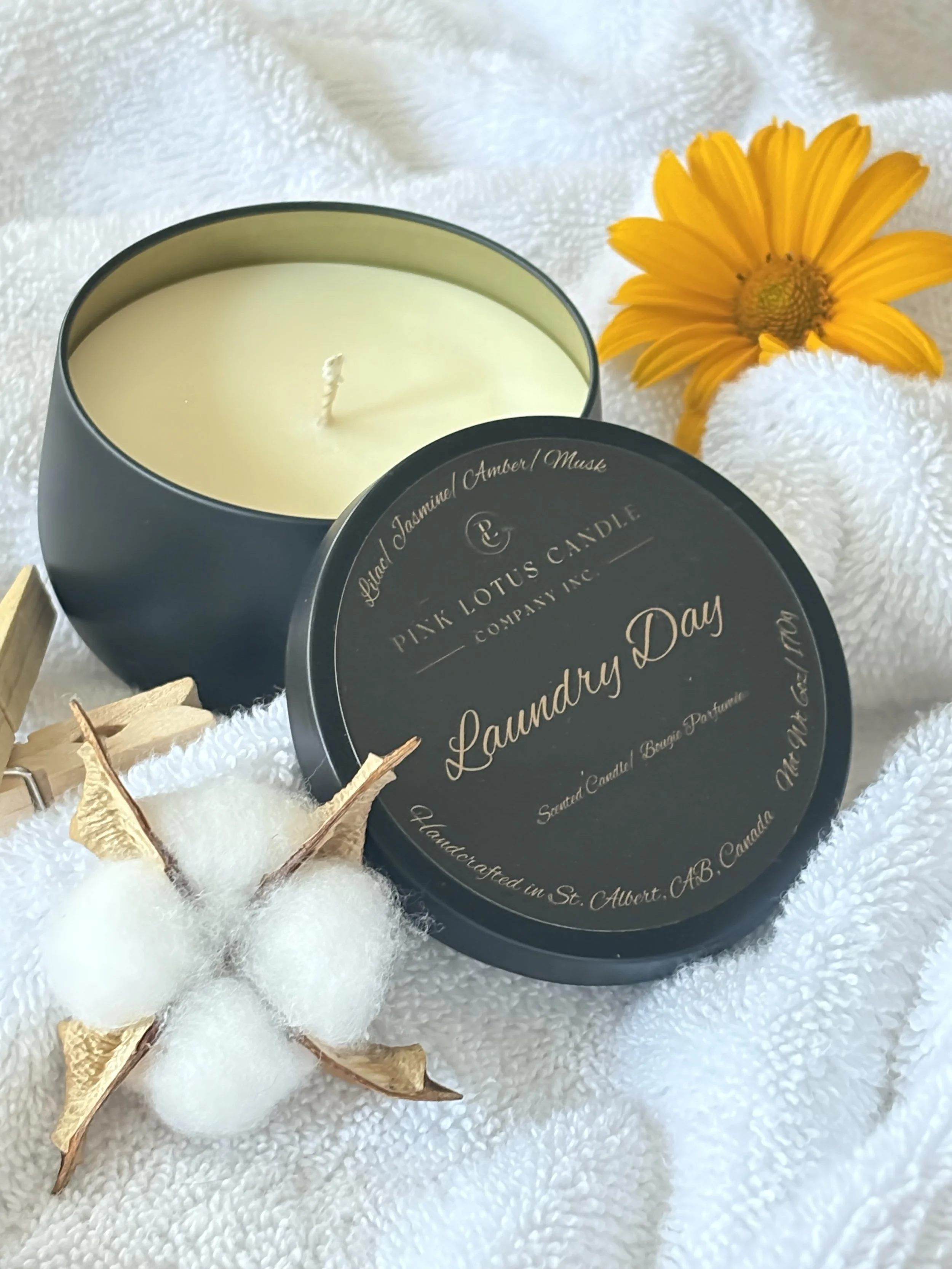 A Laundry Day scented black candle tin sitting in folded towels next to clothes pins and an orange flower
