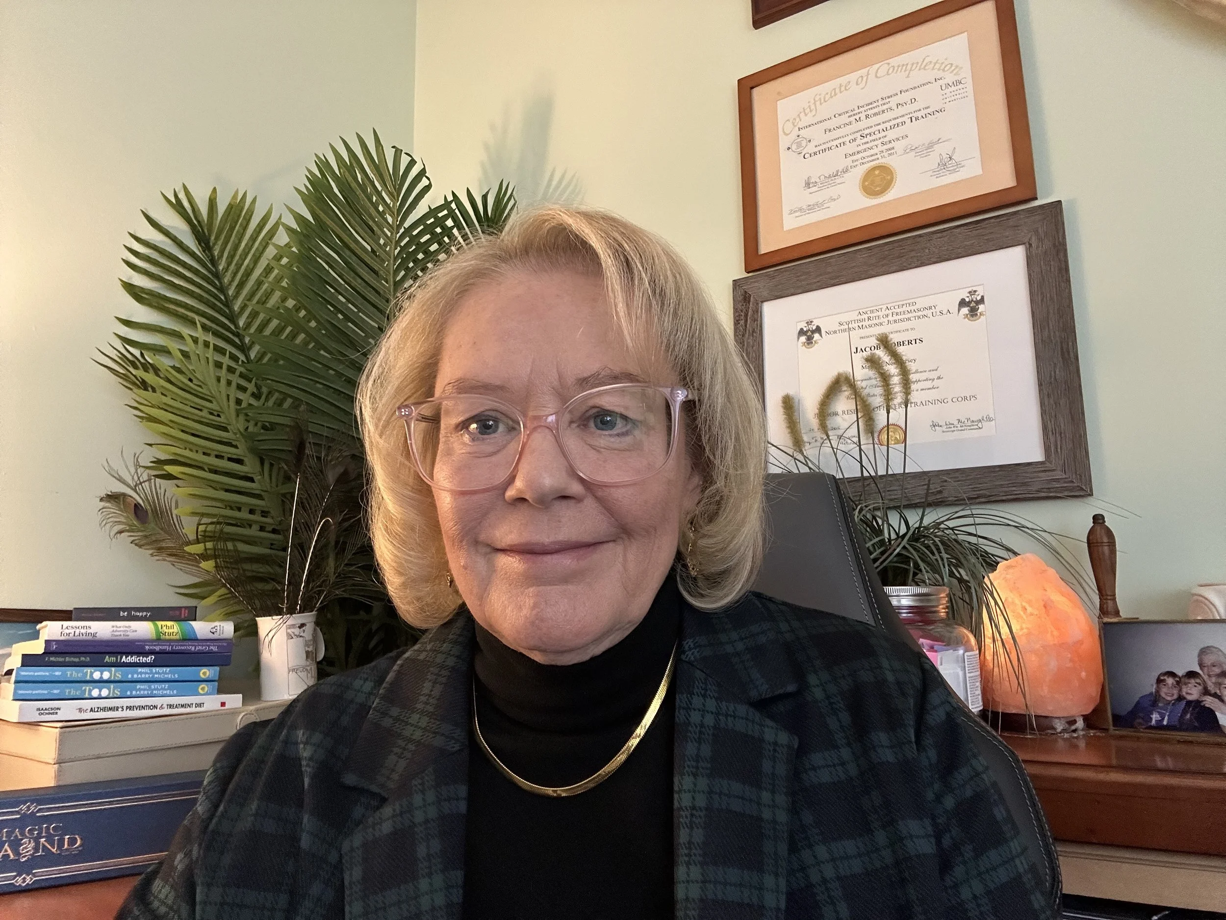 A woman with blonde hair and glasses smiling in an office. Behind her are framed certificates on a light green wall, a large green plant, a salt lamp, and a framed family photograph.