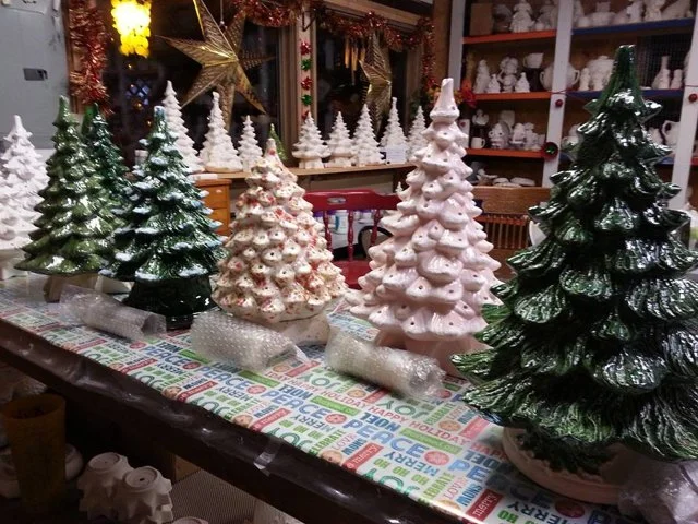 Various ceramic Christmas trees on a table with holiday-themed tablecloth and decorations in the background.