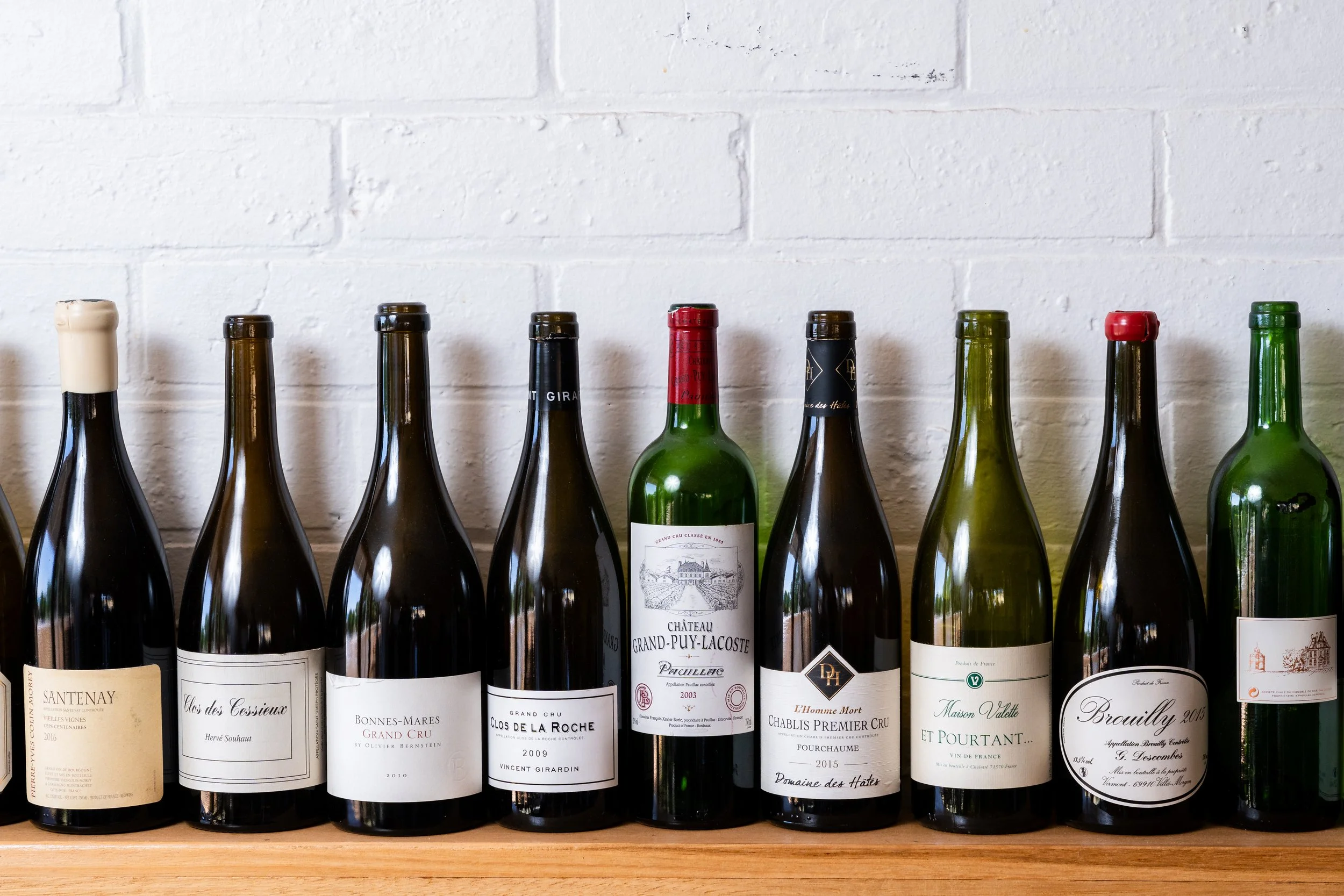 Row of various wine bottles on a wooden shelf against a white brick wall.