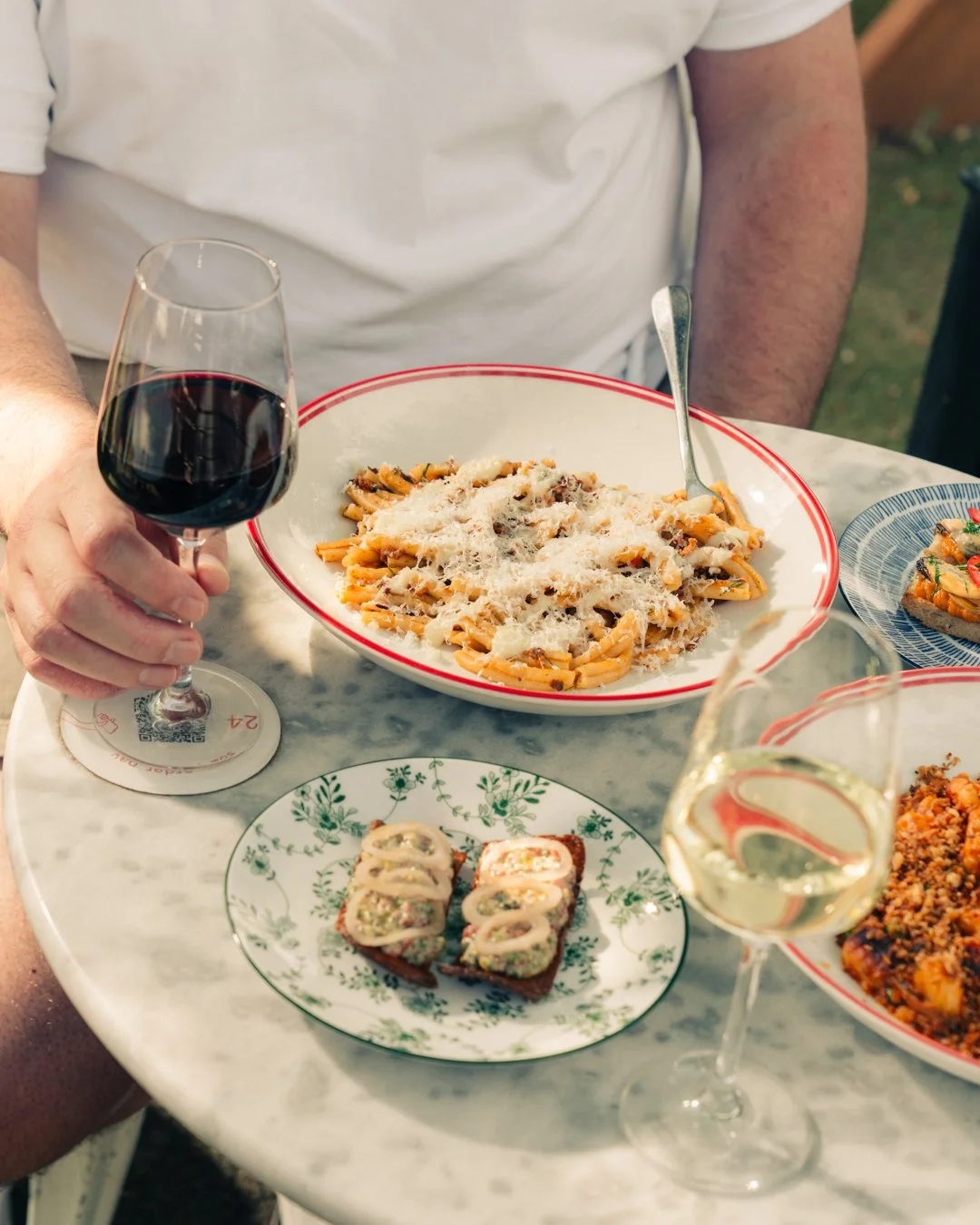 Person holding glass of red wine with plates of pasta and sushi on marble table.