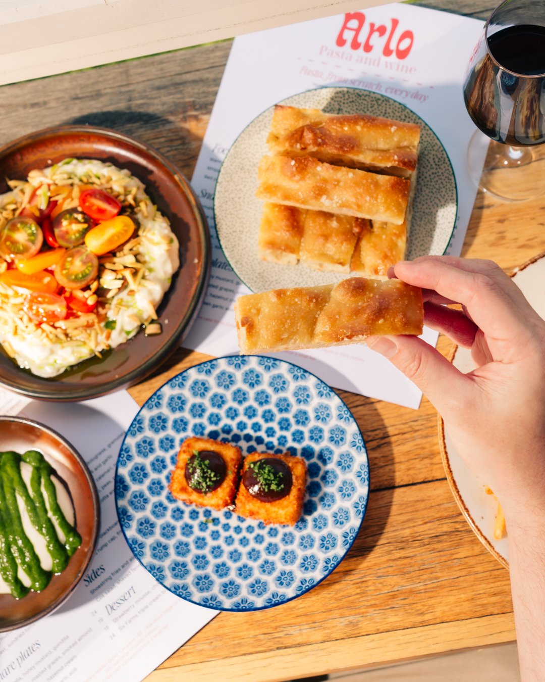 A table with various dishes including a salad with cherry tomatoes, savory fried pastries, two pieces of fried food with sauce, and sliced cucumbers, along with a glass of red wine.