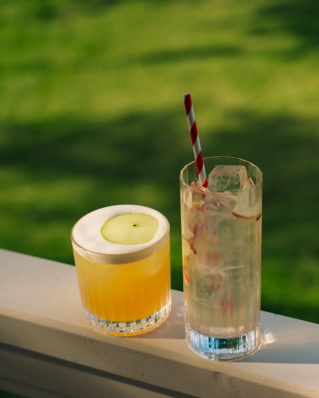 Two drinks on a ledge with a blurry green outdoor background. The left drink is a yellow cocktail with a cucumber slice on top, and the right drink is a clear beverage with ice and a red and white striped straw.