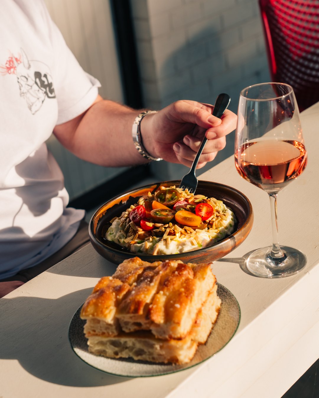 A person eating a salad with cherry tomatoes and nuts, a glass of rosé wine, and a layered cake on a white table.