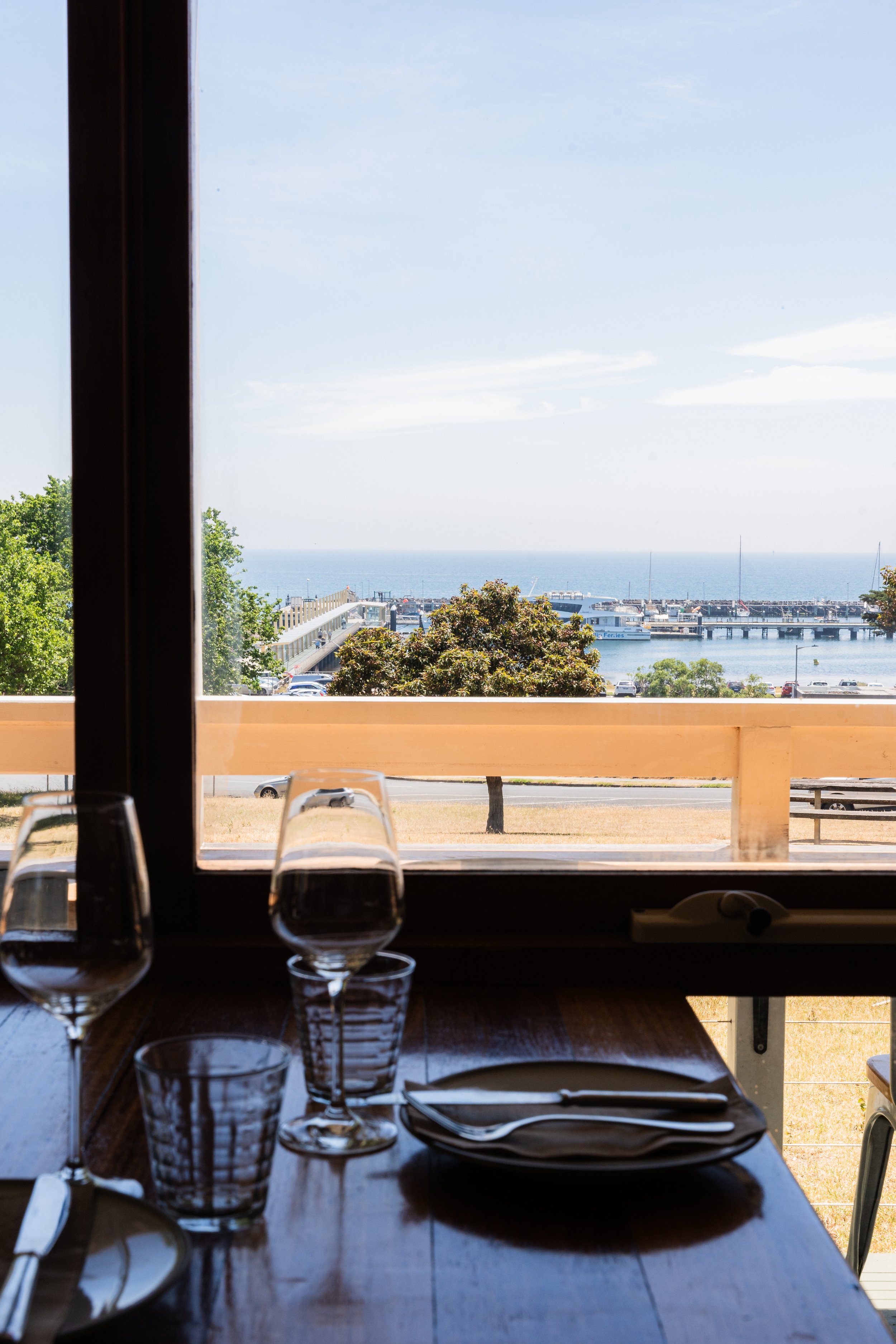 A dining table set with plates, glasses, and cutlery inside a restaurant, looking out through a window at a marina and the ocean beyond with a clear blue sky.