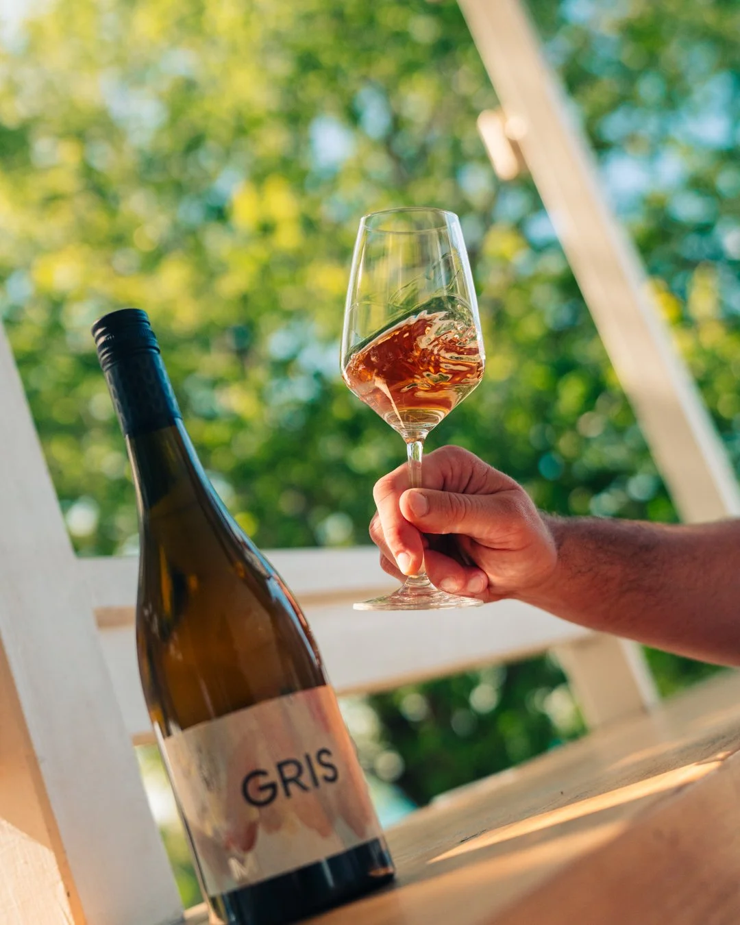 A person holds a glass of rosé wine outdoors, with a bottle of Gris wine on a wooden surface, and green trees in the background.