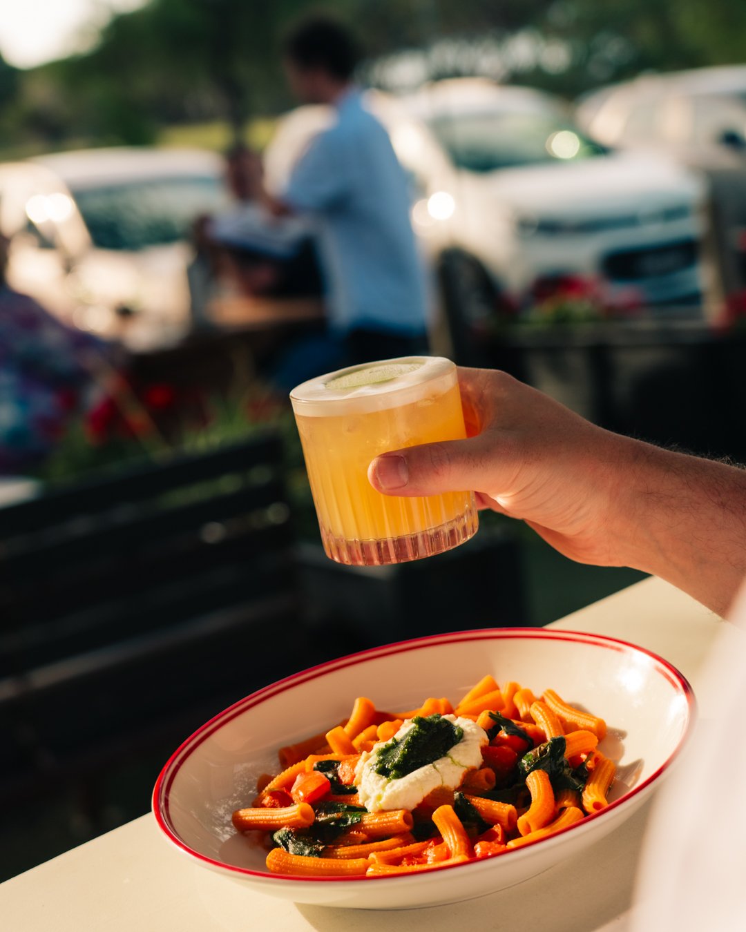 A person's hand holding a cocktail over a bowl of tomato and vodka pasta with stracciatella cheese, outdoors