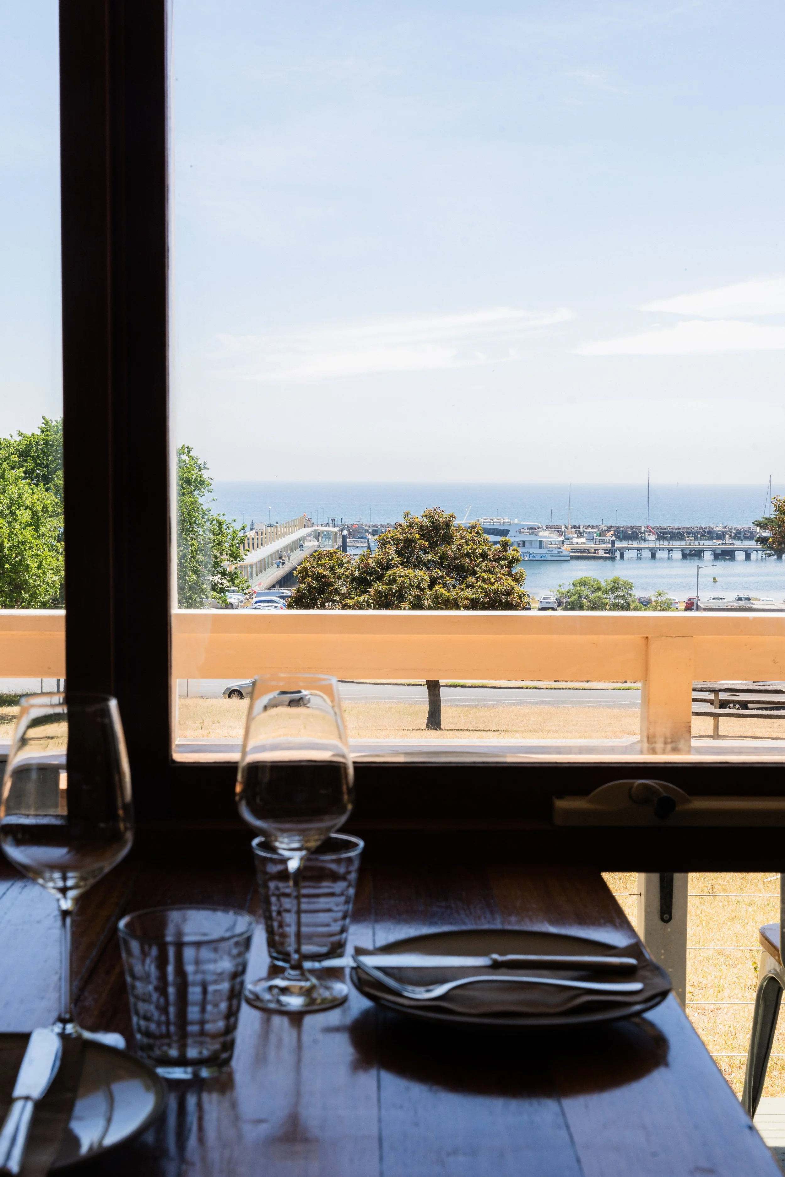 Dining table set with glasses, plates, and utensils, overlooking a harbor with boats and a pier through a large window, with trees and blue sky outside.