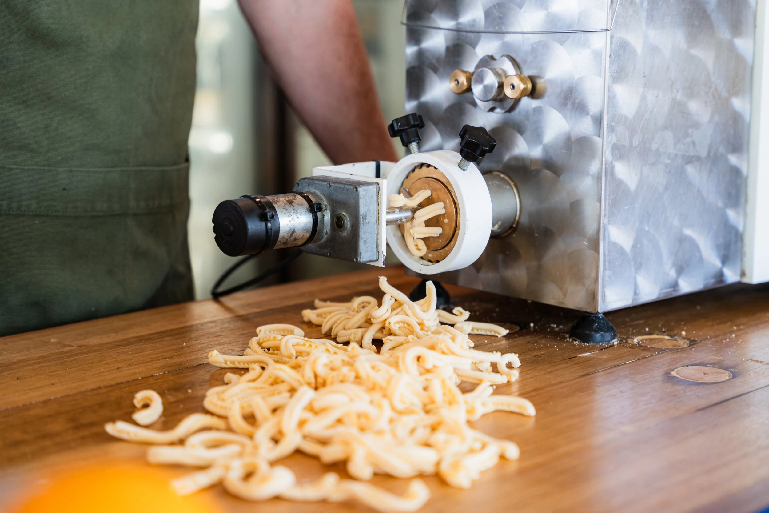 Pasta extruder machine pressing pasta onto a wooden surface with pasta spilling out of the machine.
