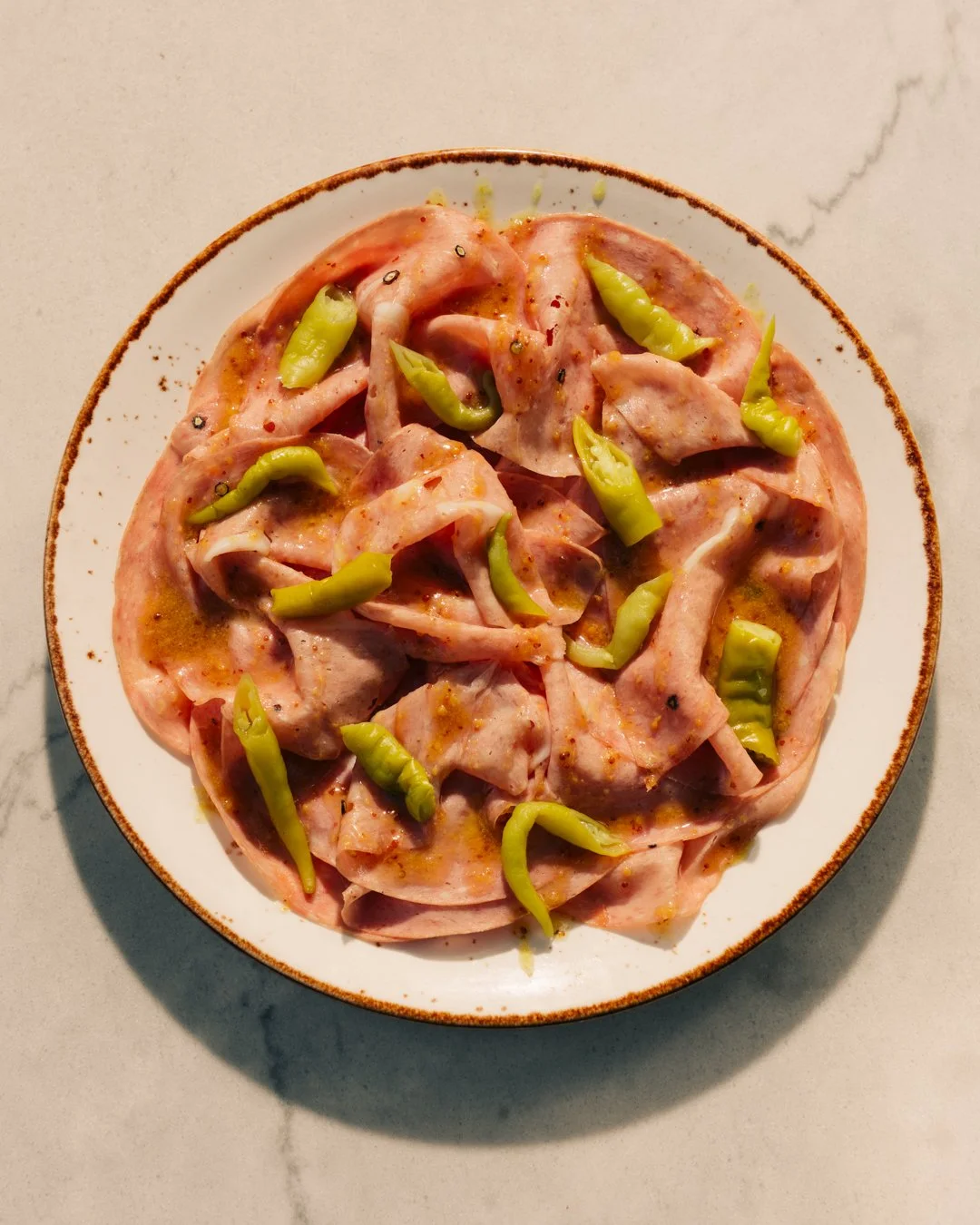 A white plate with a brown rim containing sliced ham, topped with sliced green chili peppers, on a light-colored marble surface.