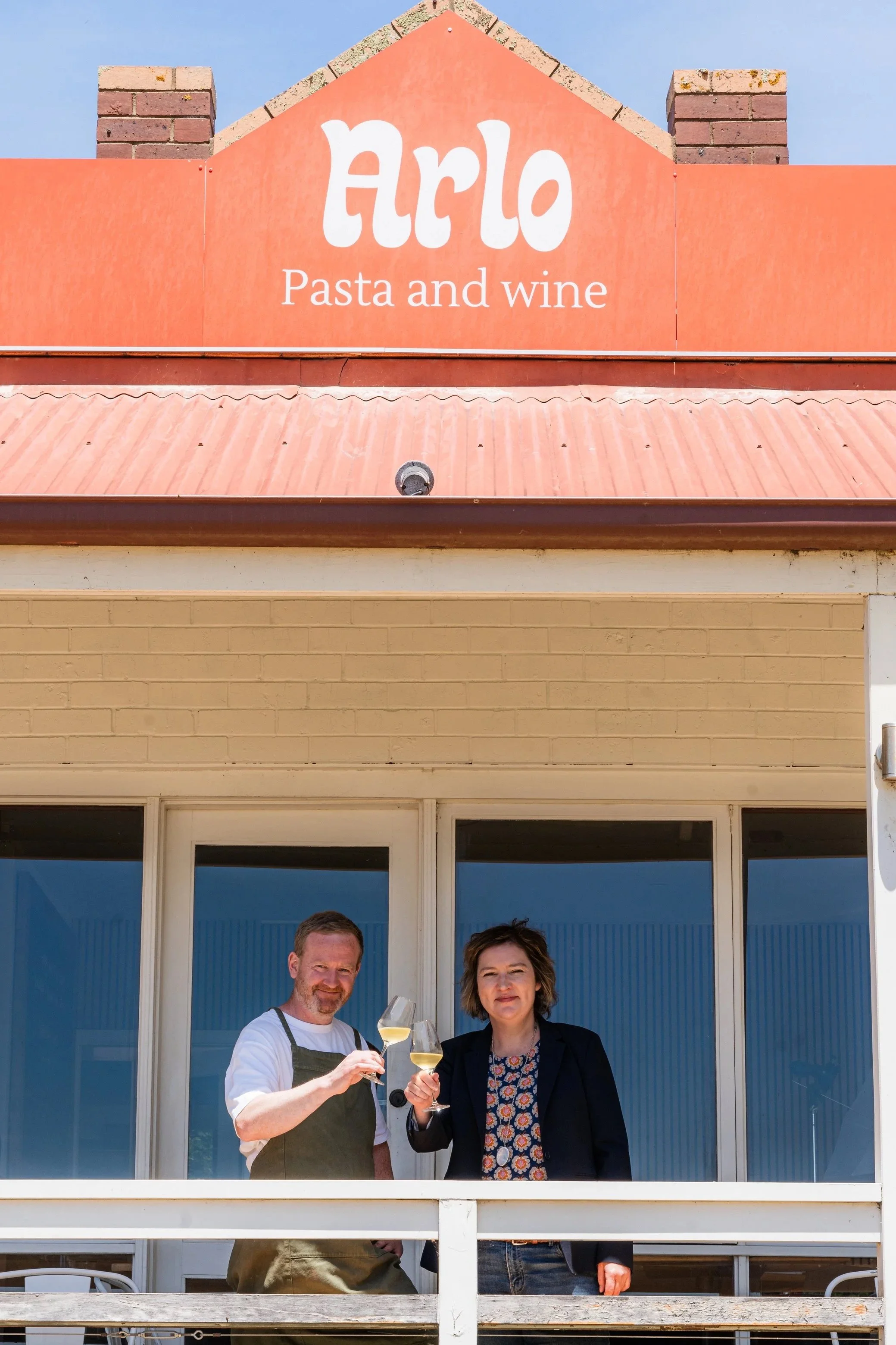 Two people holding glasses of white wine, standing on a balcony in front of large glass doors. The man is wearing a white shirt and apron, and the woman is wearing a black blazer with a patterned blouse.