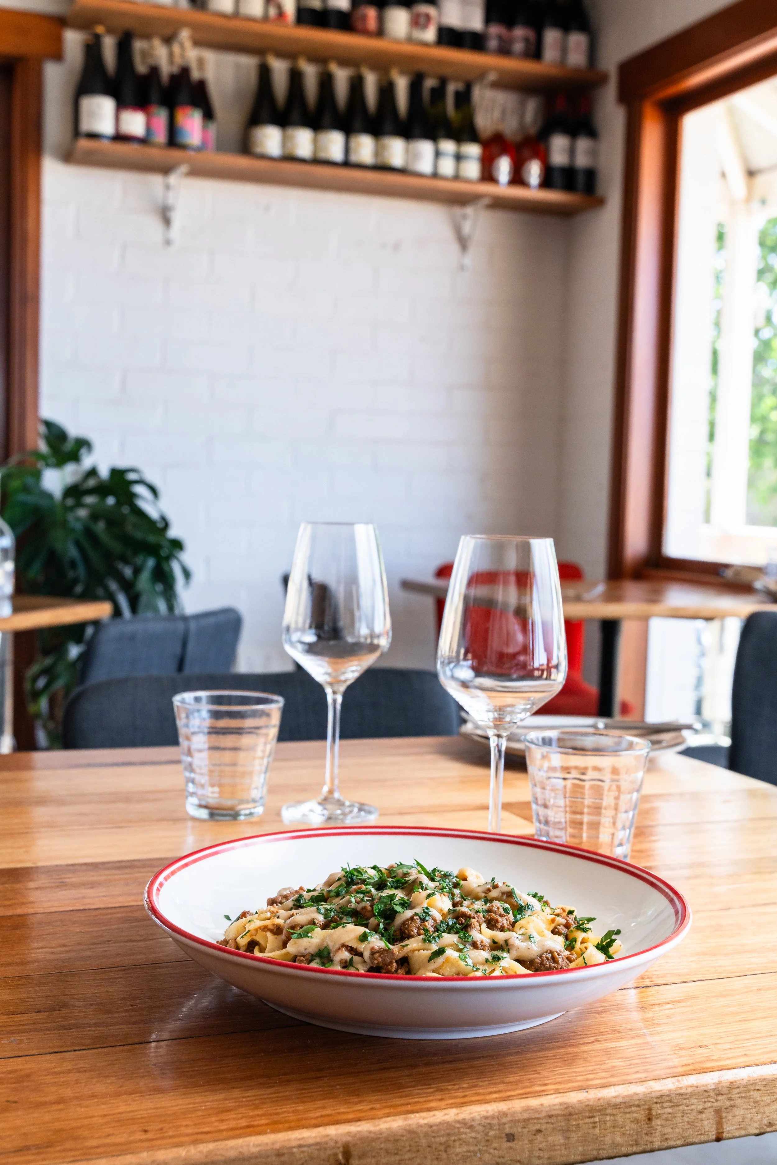 A plate of pasta with ground meat and chopped herbs on a wooden table set for dining, with wine glasses and water glasses, inside a bright restaurant with wooden accents and a window.