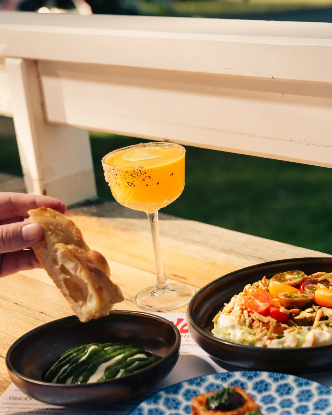 A dining table with a slice of pizza, a cucumber salad, and a colorful salad with cherry tomatoes and nuts, a yellow cocktail with lemon, and a hand holding a slice of pizza in front of the drinks.