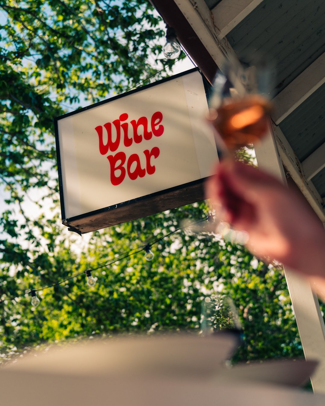 Sign for a wine bar with a person holding a glass of rosé wine in the foreground, outdoors on a sunny day with green trees in the background.