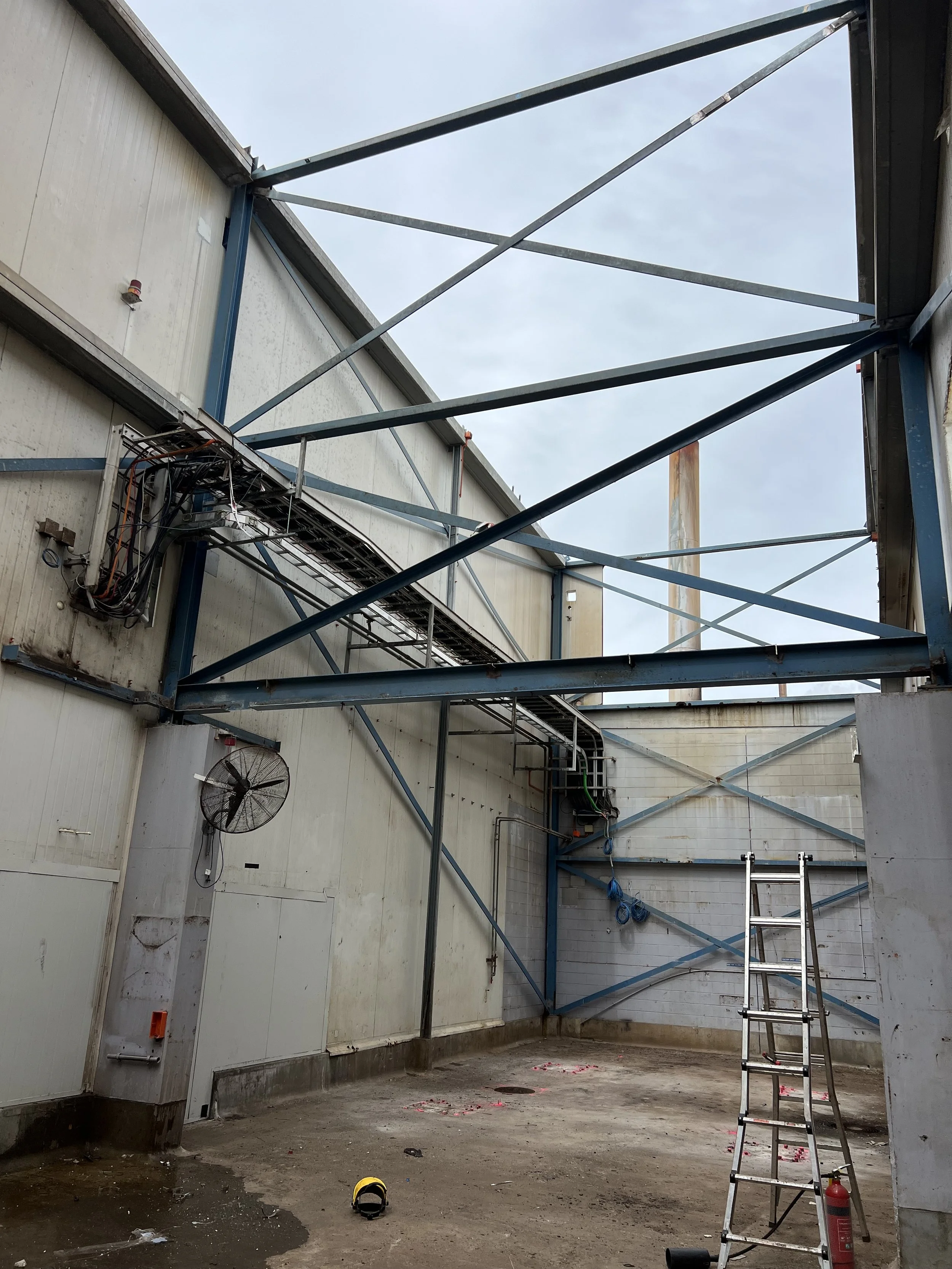 Construction site with blue steel framing, a ladder, a fire extinguisher, and a wall-mounted fan inside an industrial building under construction.
