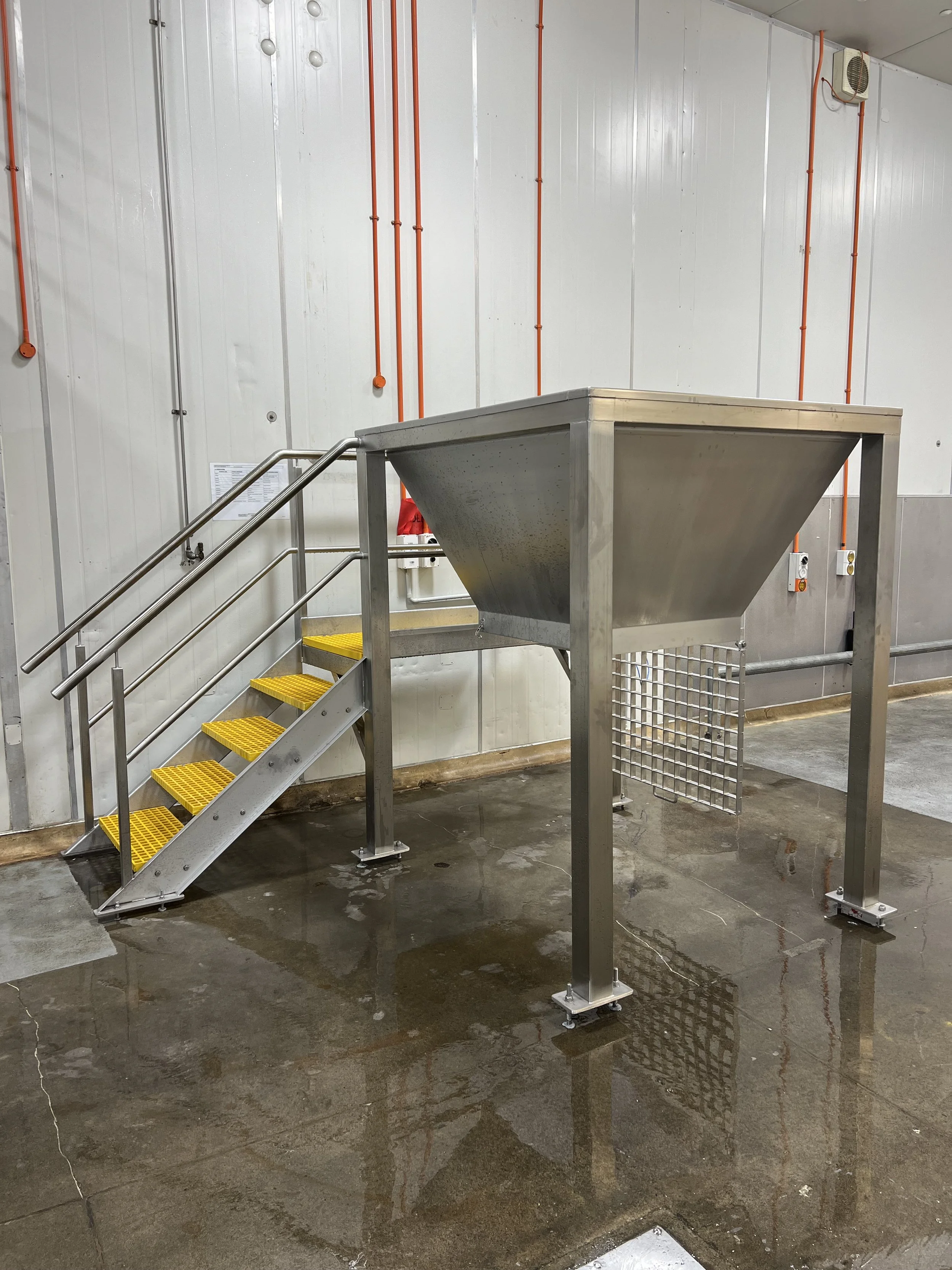 Industrial metal staircase leading to a large metal hopper in a warehouse or factory with wet and stained concrete floor.
