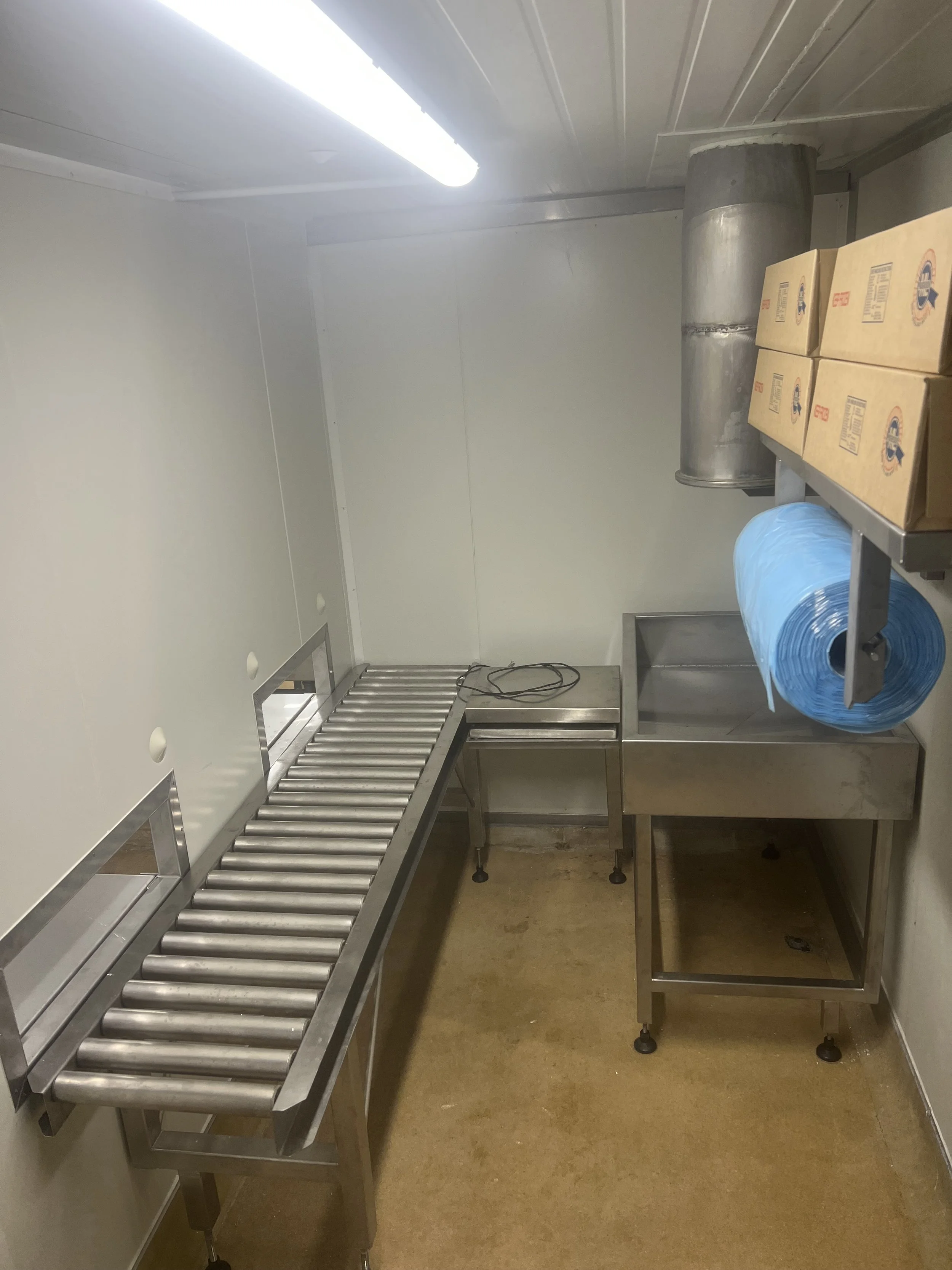 A small industrial kitchen or storage room with a metal sink, a blue roll of plastic trash bags, and tarped boxes on metal shelving. Fluorescent light on ceiling, metal ventilation duct, and beige floor.