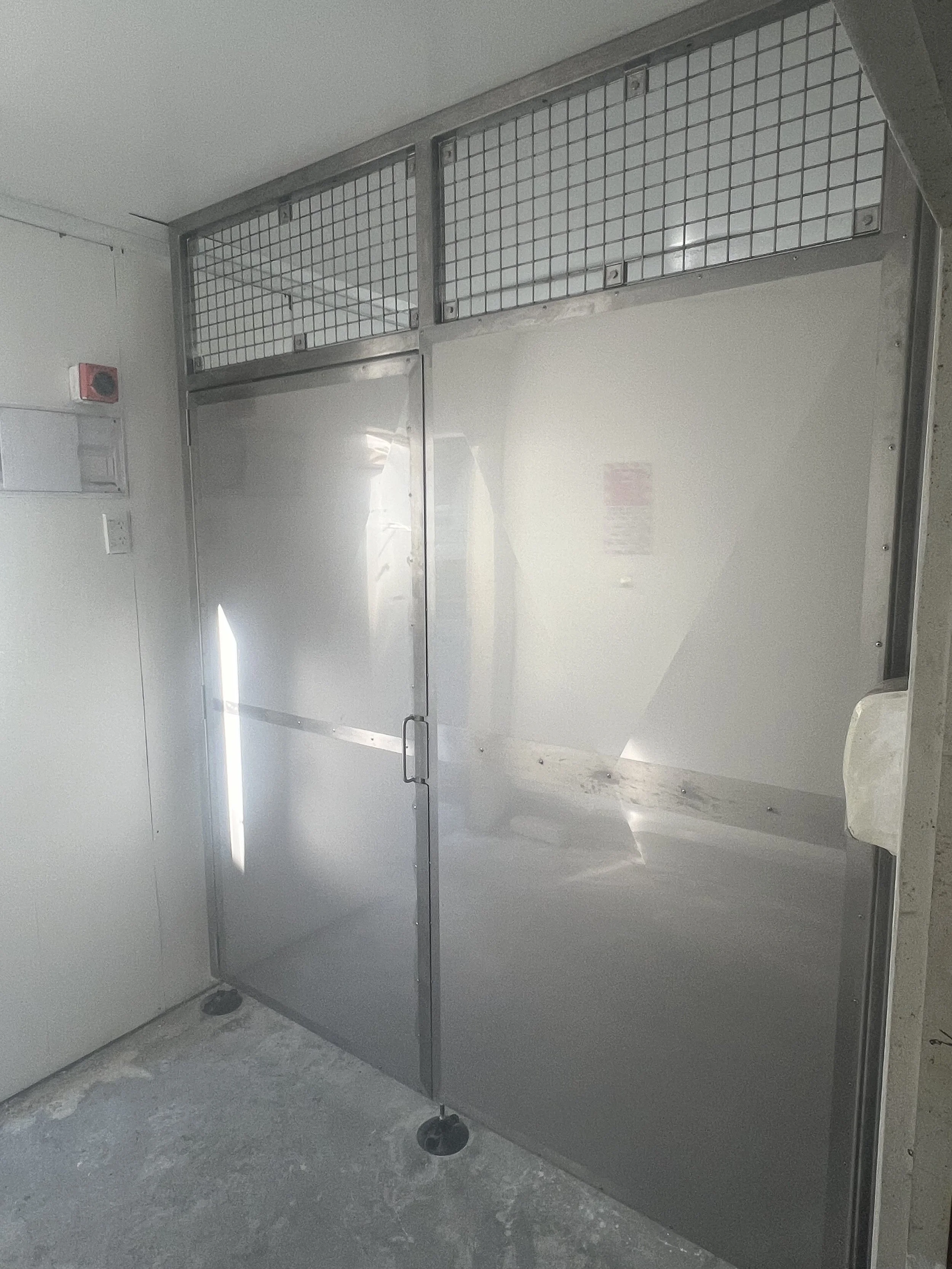 Steel door with a handle and a mesh section at the top, installed in a wall with a small red alarm or sensor on the adjacent wall.