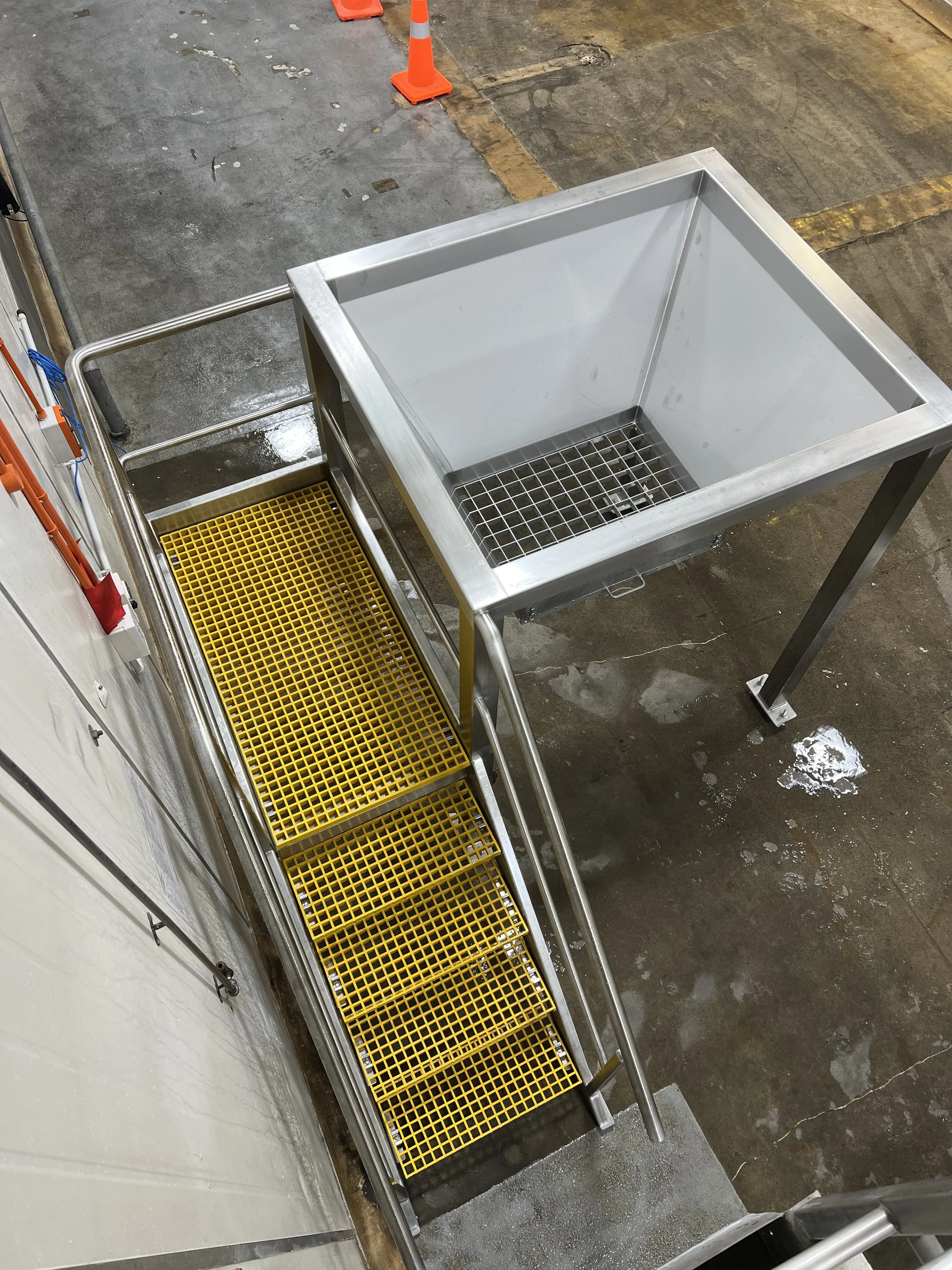 Industrial workspace with a stainless steel sink and a yellow grated platform with stairs, located on a concrete floor with orange safety cones nearby.