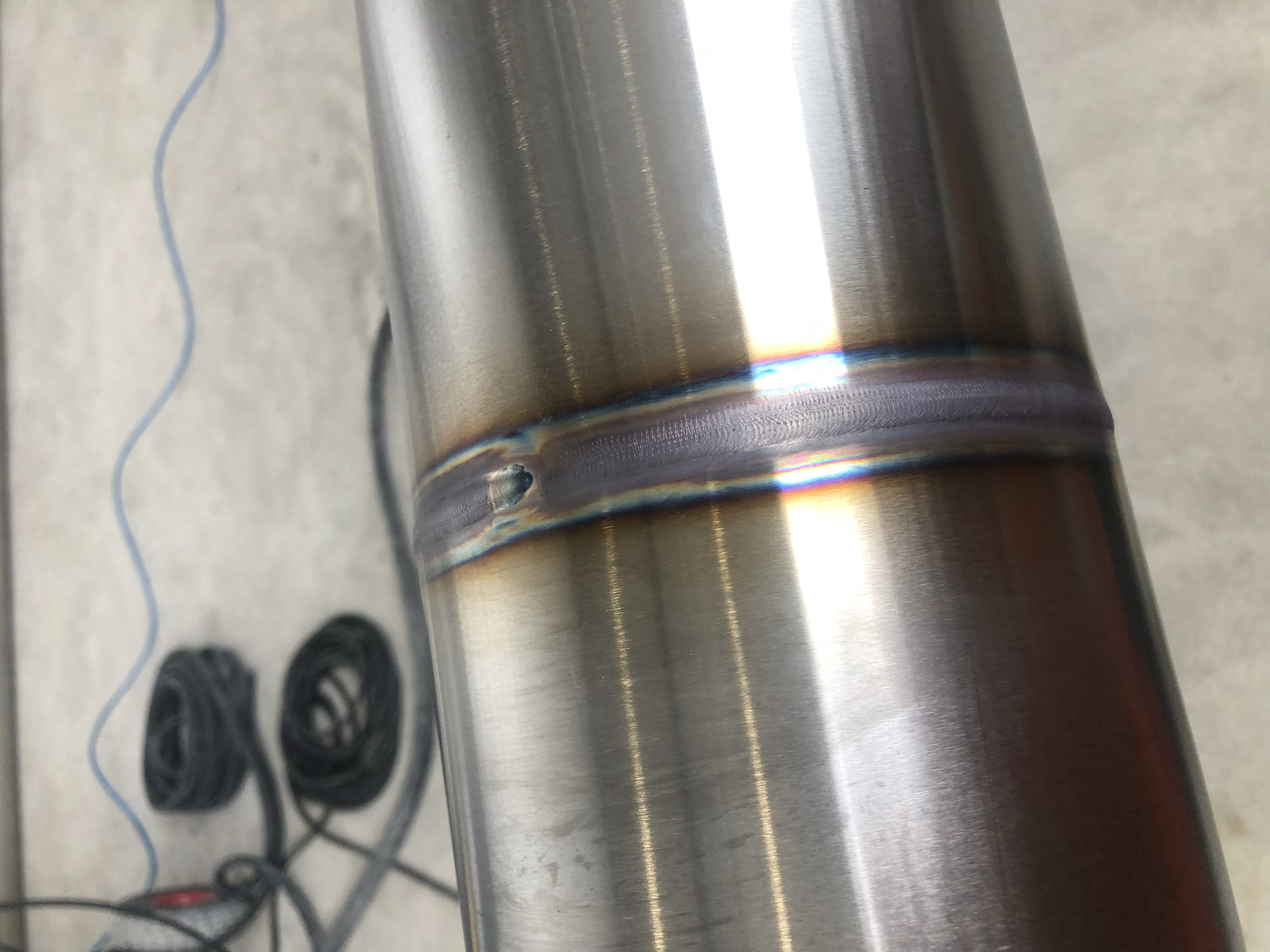 Close-up of a metallic pipe with a weld seam, showing heat discoloration and a small hole near the weld. In the background, black cords and a blue wire are visible on the concrete floor.