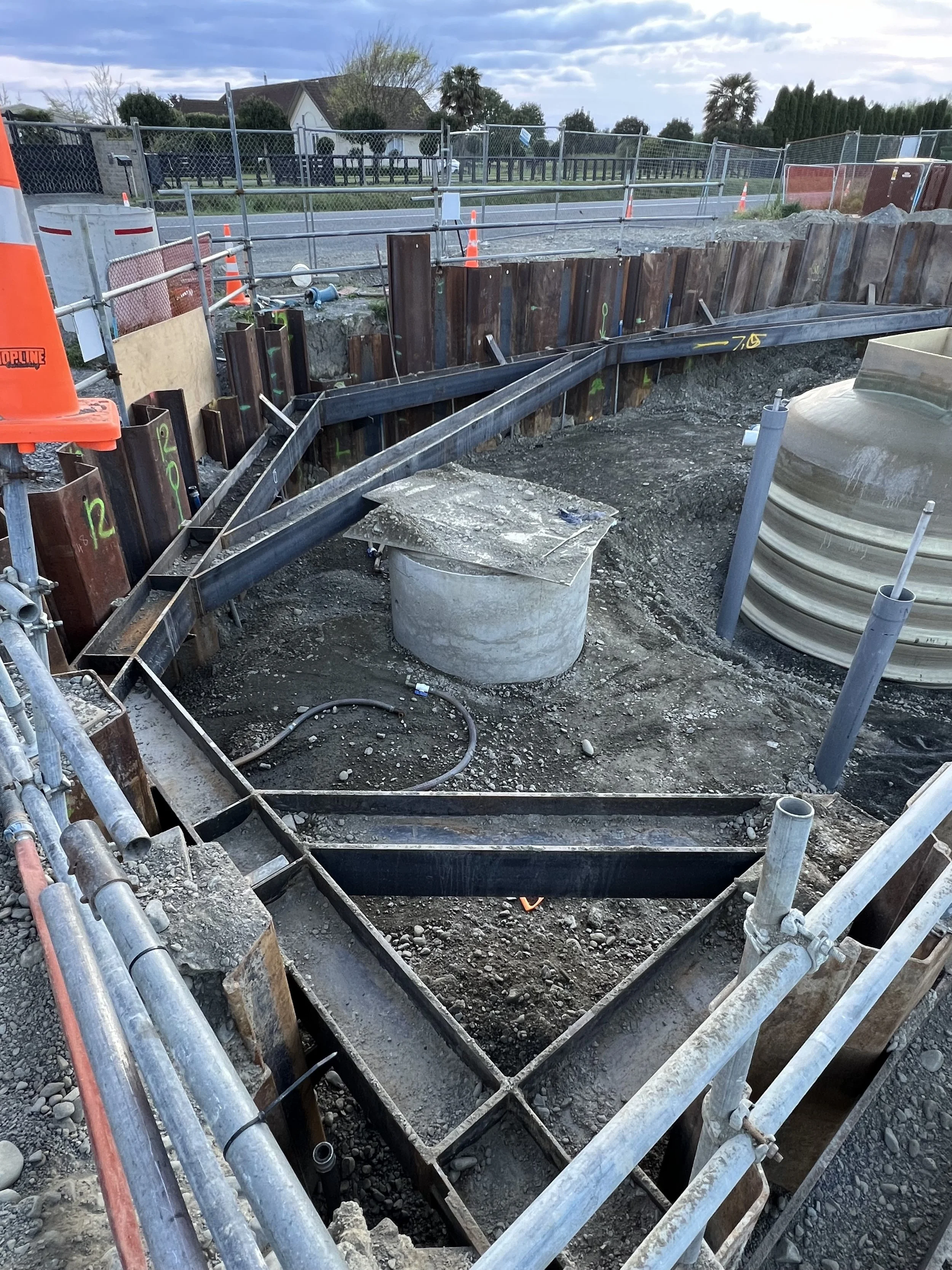 Construction site with foundation work, metal reinforcements, and pipes, surrounded by a temporary fence and orange safety barriers.