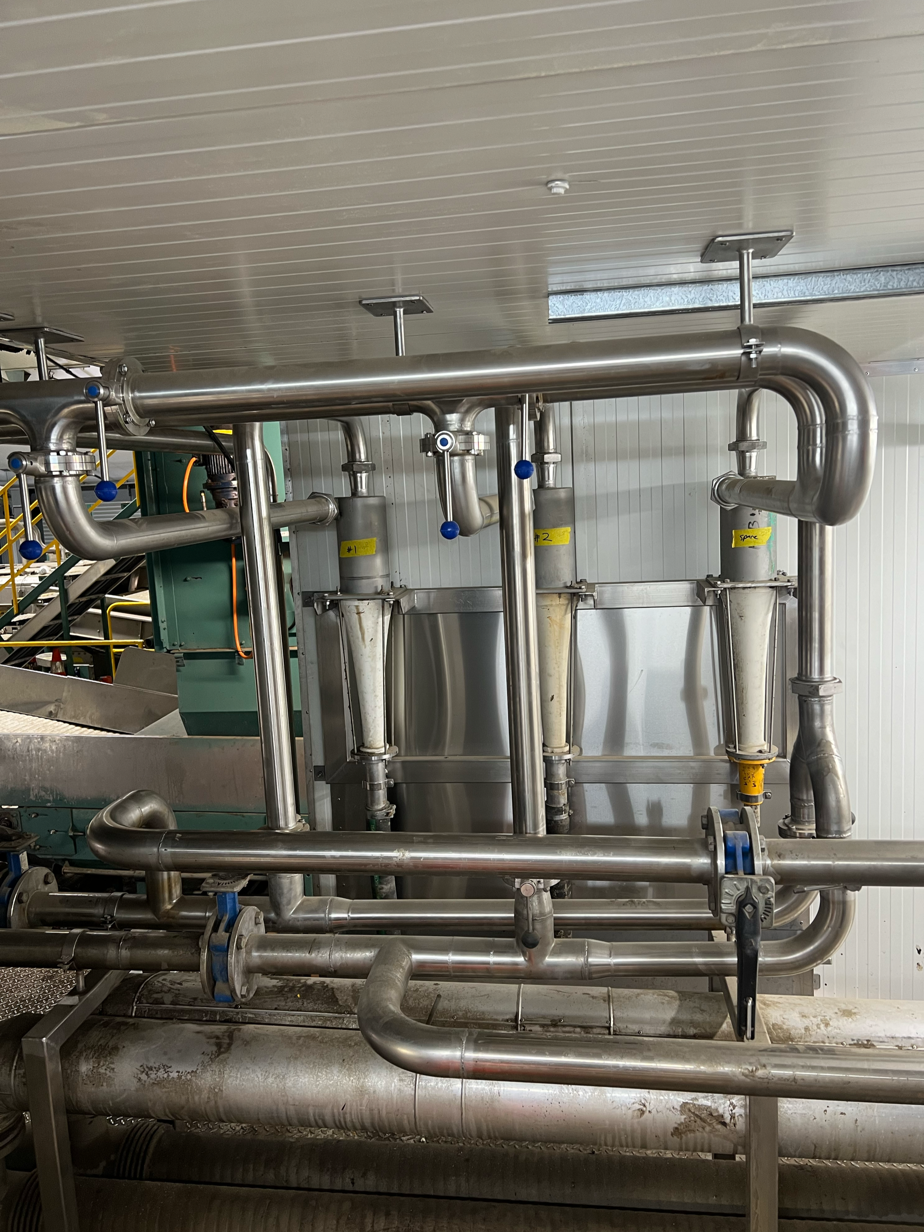 Industrial stainless steel pipes and valves with three cone-shaped containers labeled 1, 2, and spare, in a factory setting.