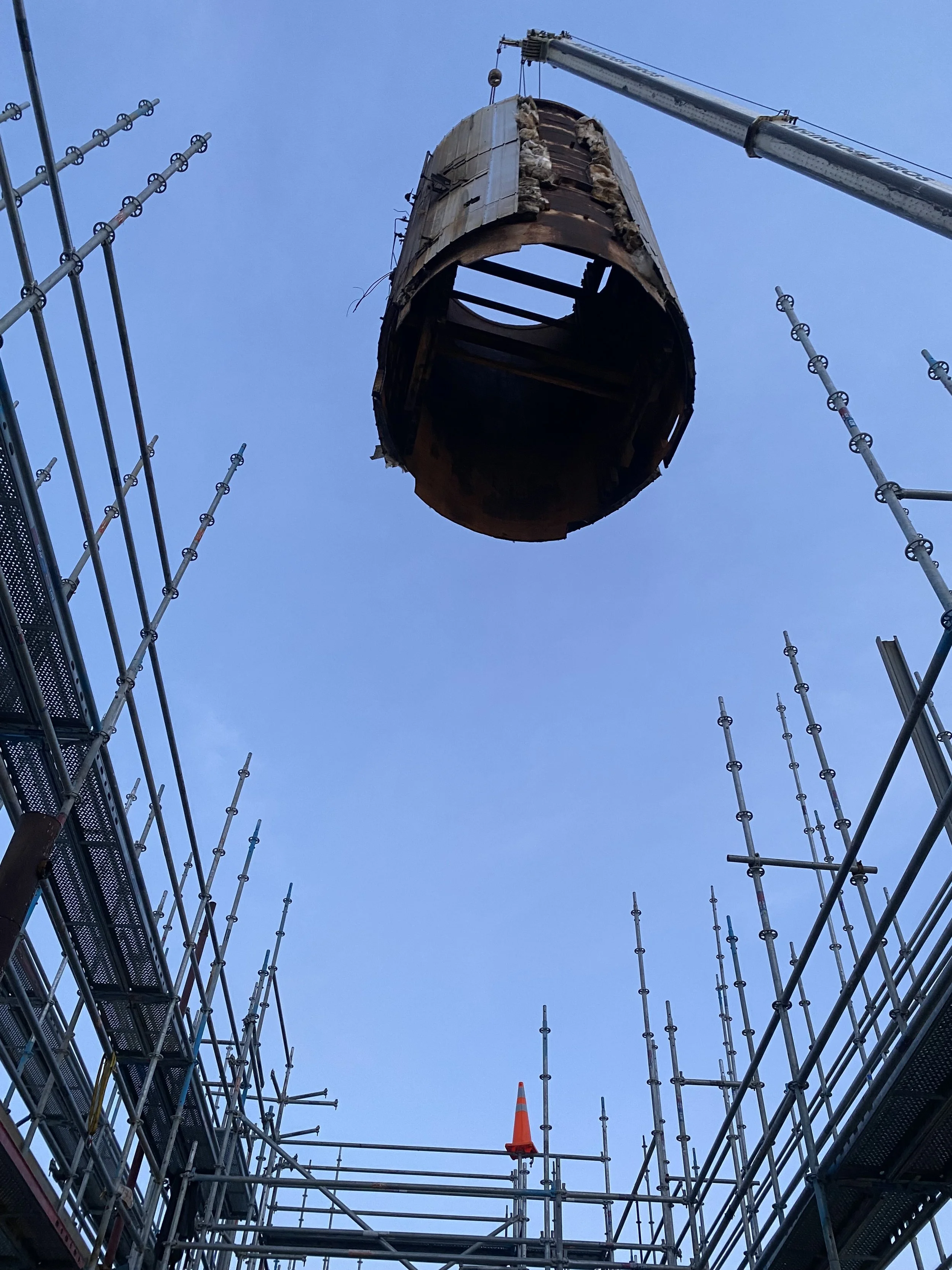 Construction site with a large, hollow, cylindrical metal structure being lifted by a crane, surrounded by scaffolding and an orange safety cone.