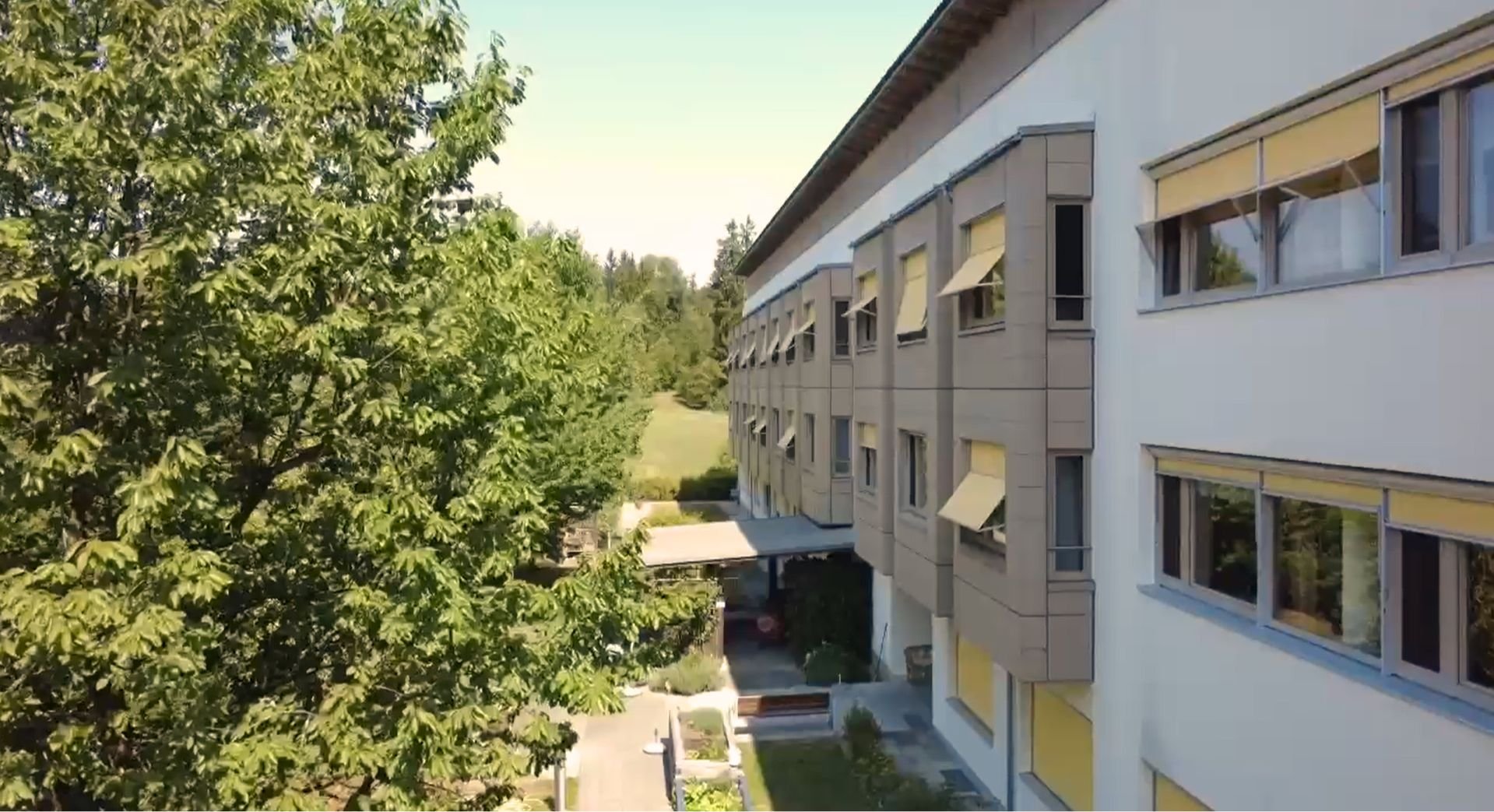 Das Gebäude des Sozialzentrums Sonnleiten mit mehreren geöffneten Fensterläden und Sonnenschutzmarkisen, umgeben von grünen Bäumen an einem sonnigen Tag.
