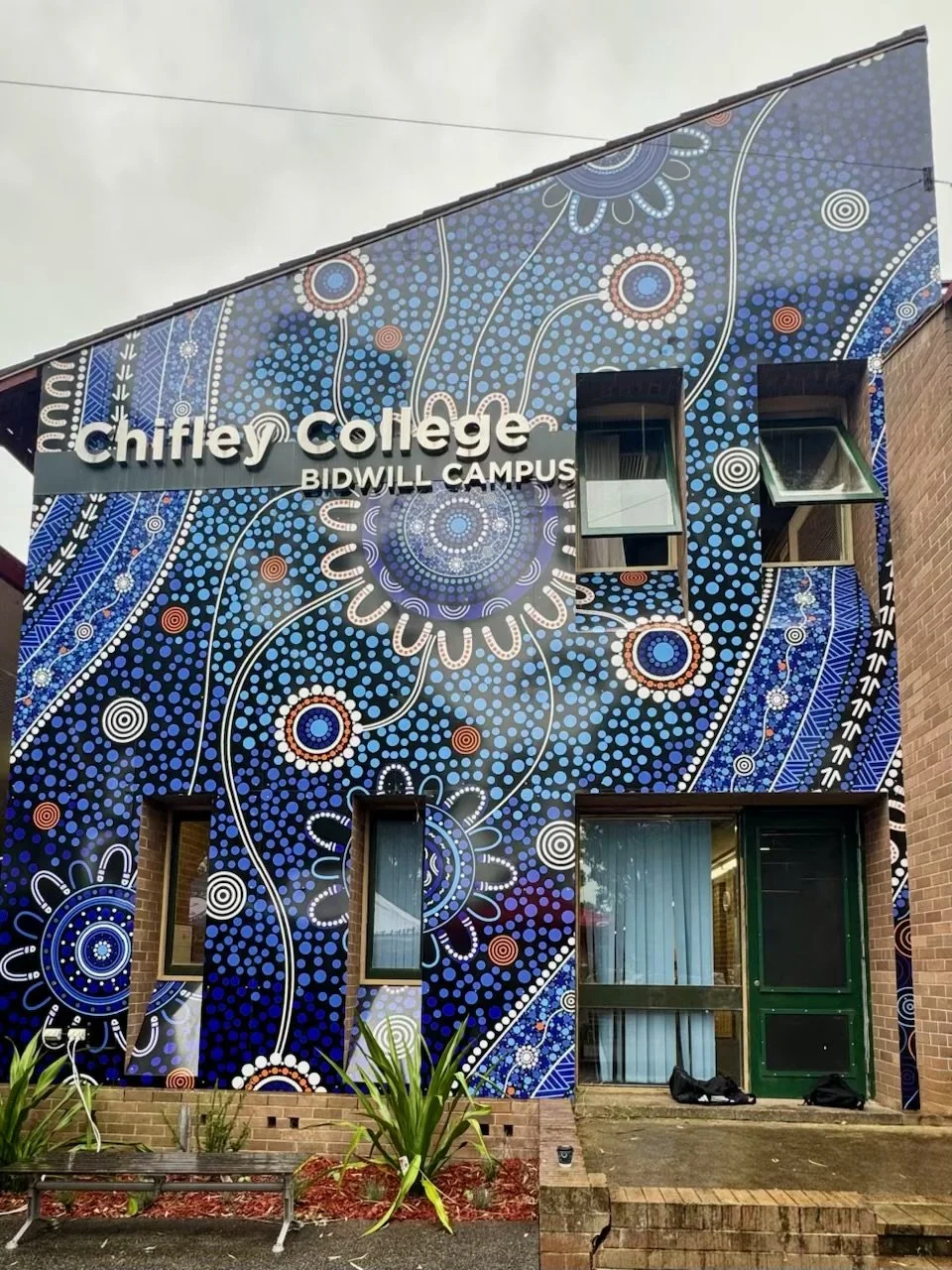 Colorful mural on the exterior of a building at Chifley College Bidwill Campus, featuring blue, white, red, and orange dot patterns and swirls, with windows and a green door.