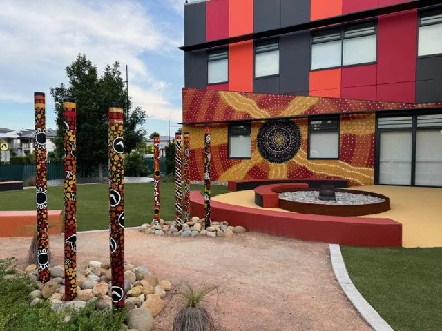 Colorful outdoor art installation featuring decorated poles with circular patterns, set against a red and yellow mural on a building.