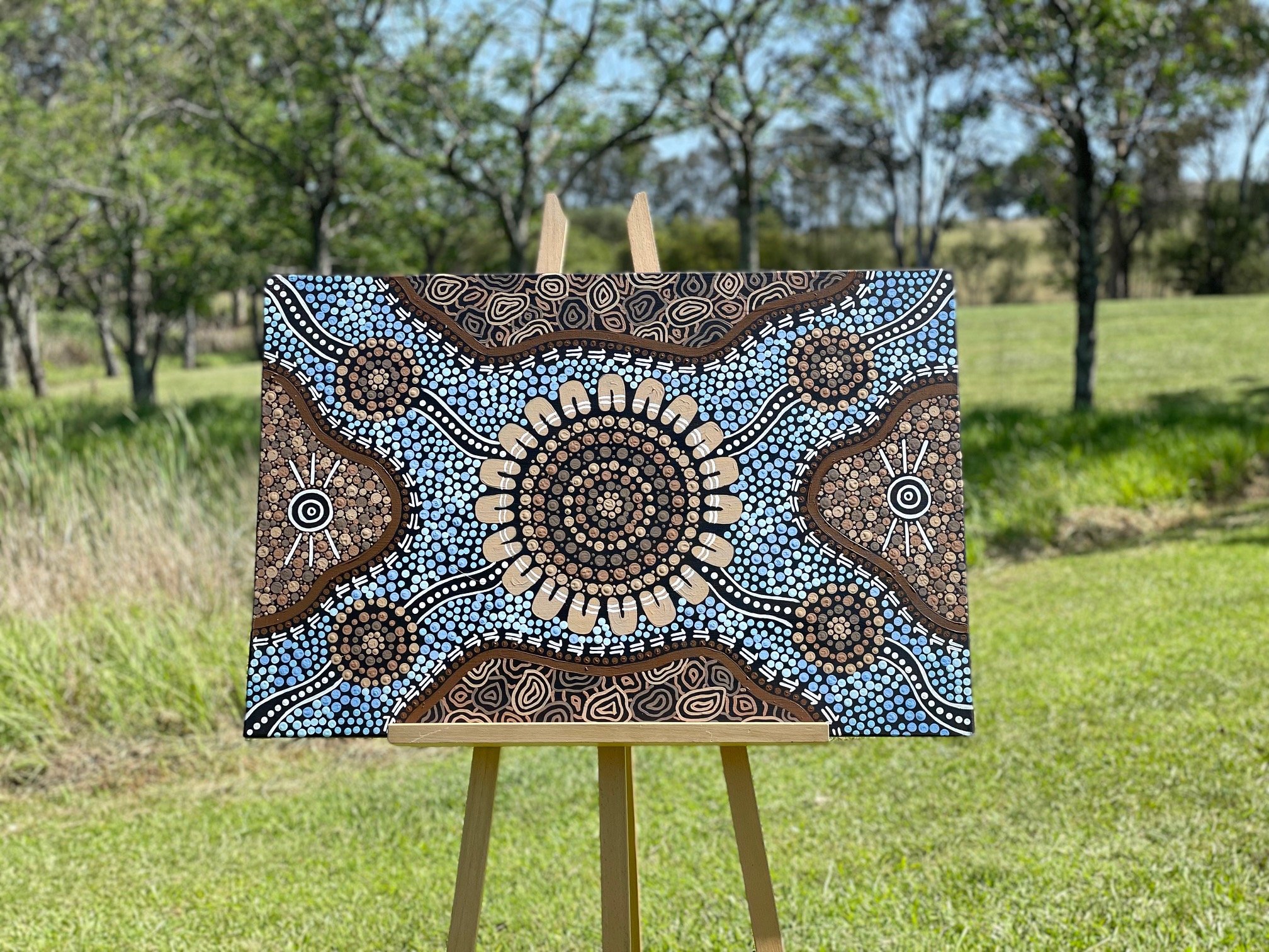 Painted canvas with Aboriginal-style dot art depicting circular and wavy patterns in shades of black, brown, beige, white, and blue, mounted on a wooden easel outdoors in a grassy area with trees in the background.
