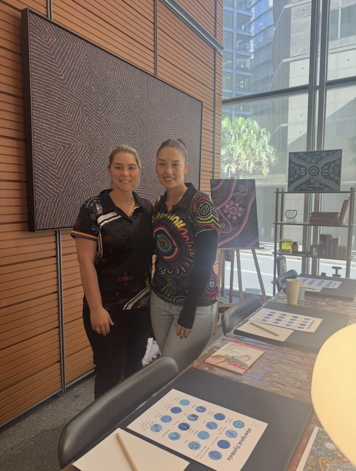 Two women standing together in an art gallery or studio, surrounded by paintings and art supplies, with a large computer monitor and a glass window showing city buildings and greenery.