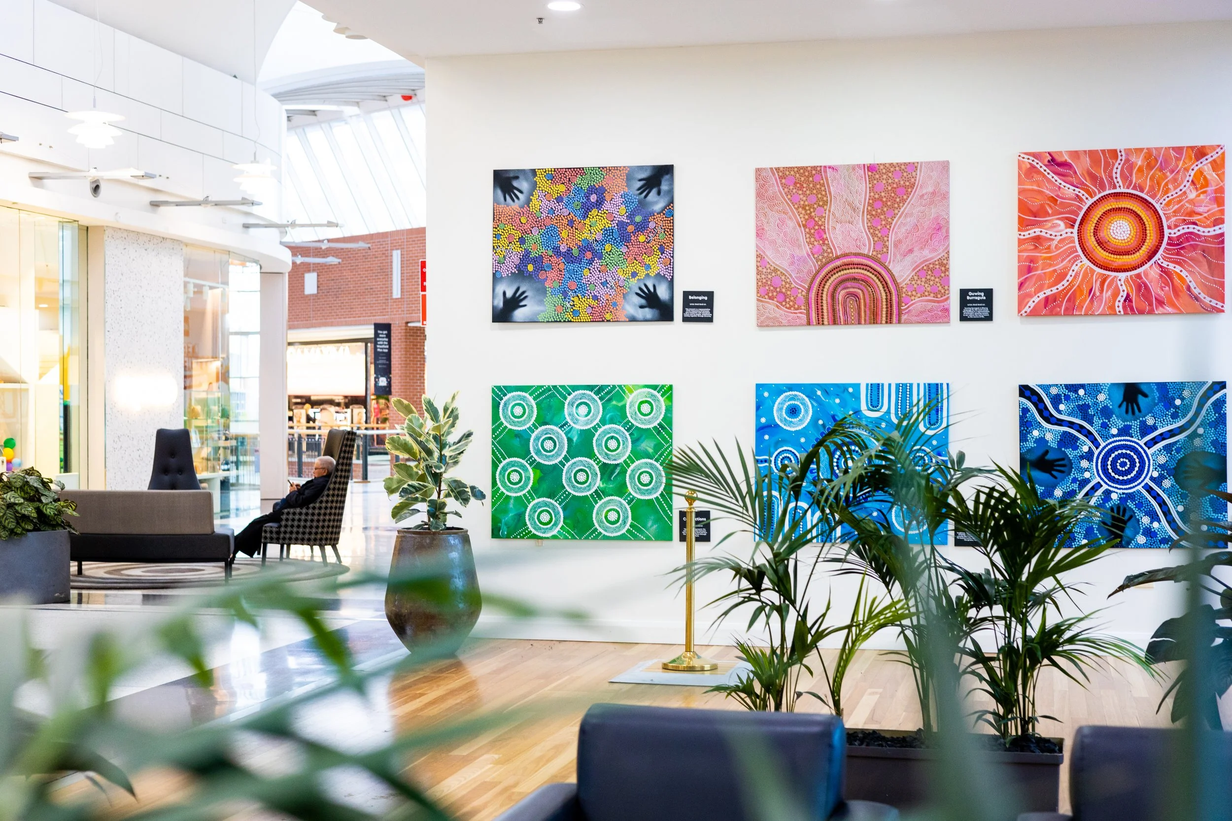 Art gallery with eight colorful paintings on white wall, plants in foreground, and seating area with a person sitting in a high-backed chair.