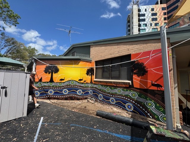 Mural painting of a sunset landscape with trees, animals, and abstract patterns on the side of a brick building.