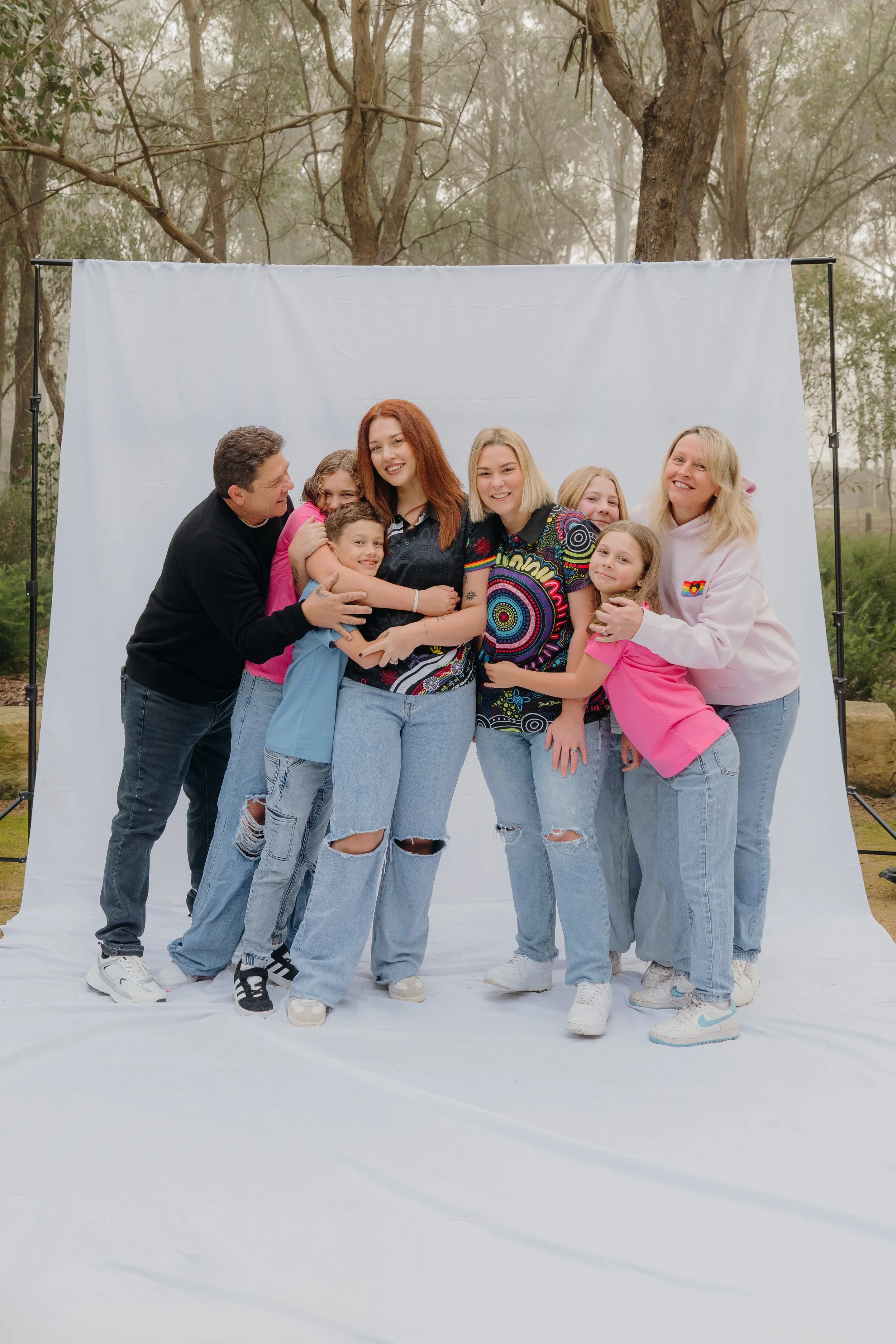 Group of people hugging and smiling in front of a white photo backdrop outdoors with trees in the background.