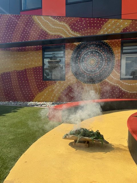 A colorful mural painted on the side of a building with windows. In the foreground, a yellow and red surface has a small heap of green leaves and steam rising above it.