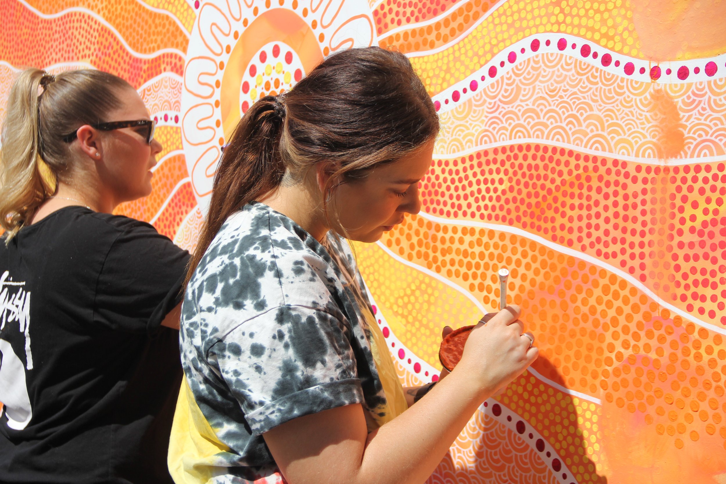 Two women creating a colorful dot painting on a large orange and yellow abstract mural. One woman is wearing sunglasses and a black shirt, the other is focused on painting.