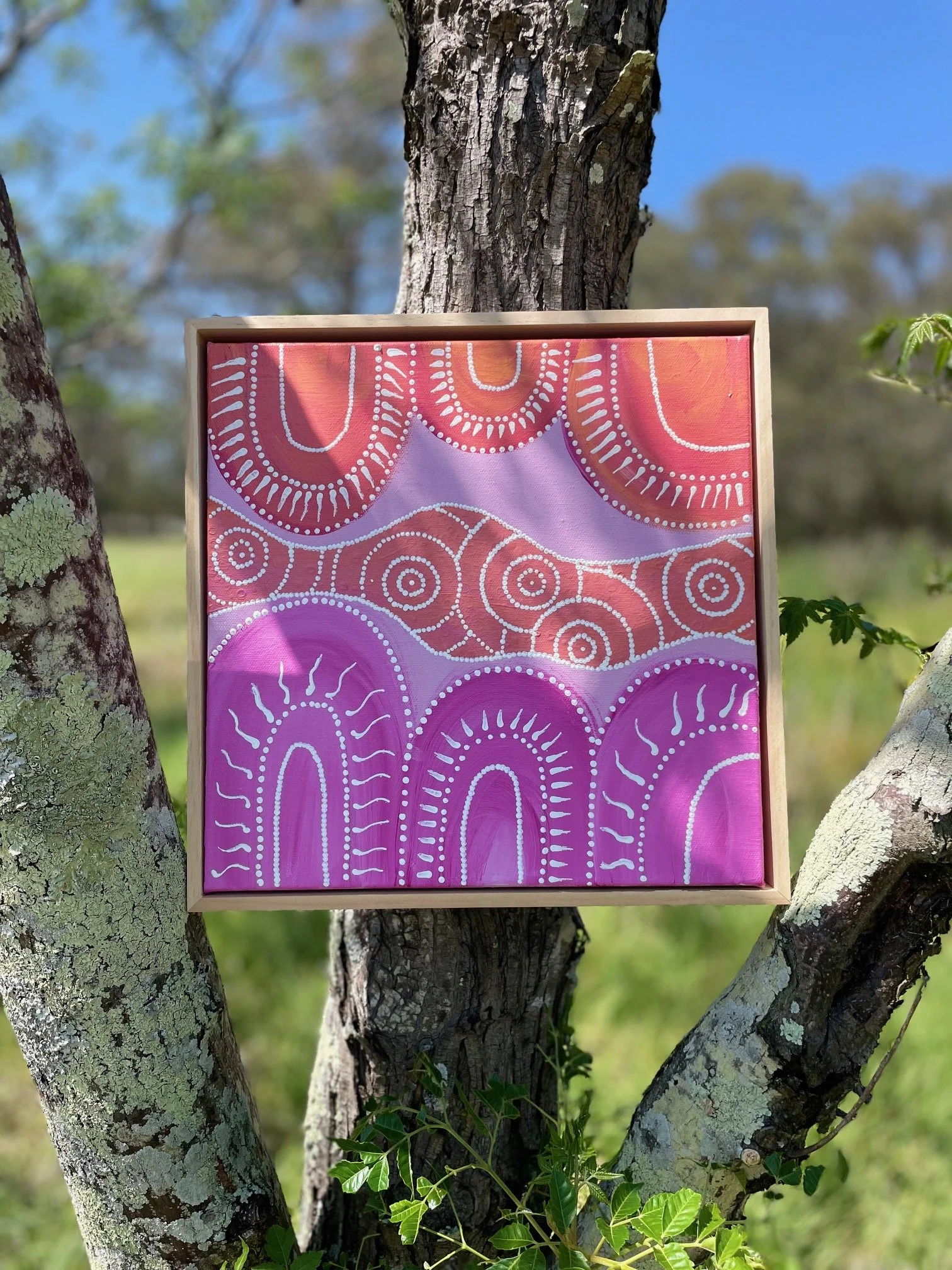 A colorful abstract painting with purple, pink, and orange hues, featuring white dotted and curved line patterns, hanging between two tree trunks outdoors.