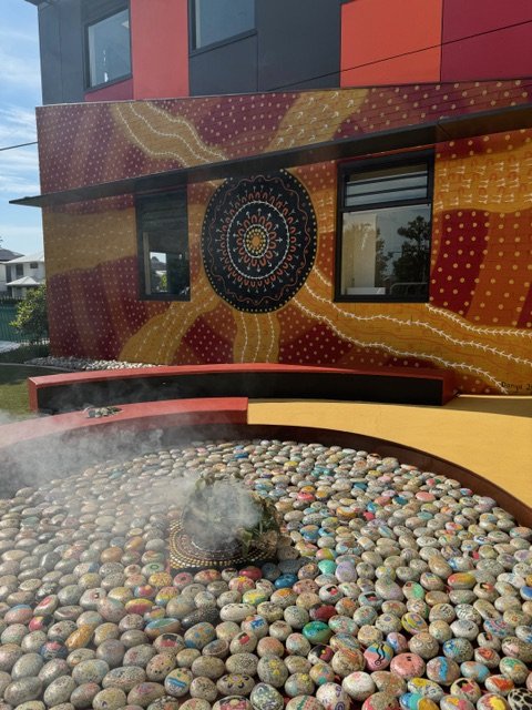 Colorful building exterior with a vibrant mural, two windows, and a water feature filled with smooth multicolored stones with steam rising from it.