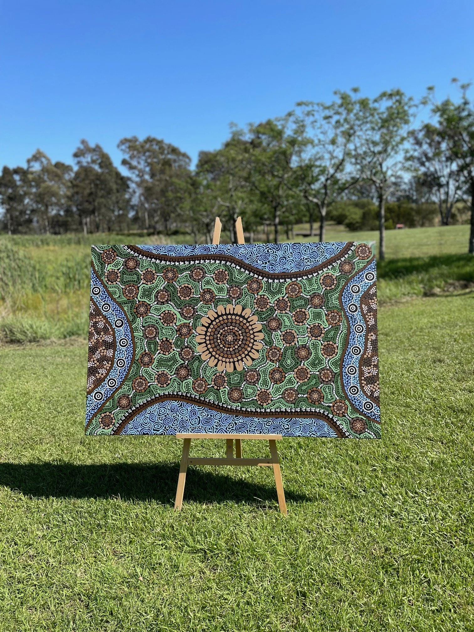 Painted canvas with colorful Aboriginal dot art on an easel outdoors in a grassy field with trees and a blue sky in the background.