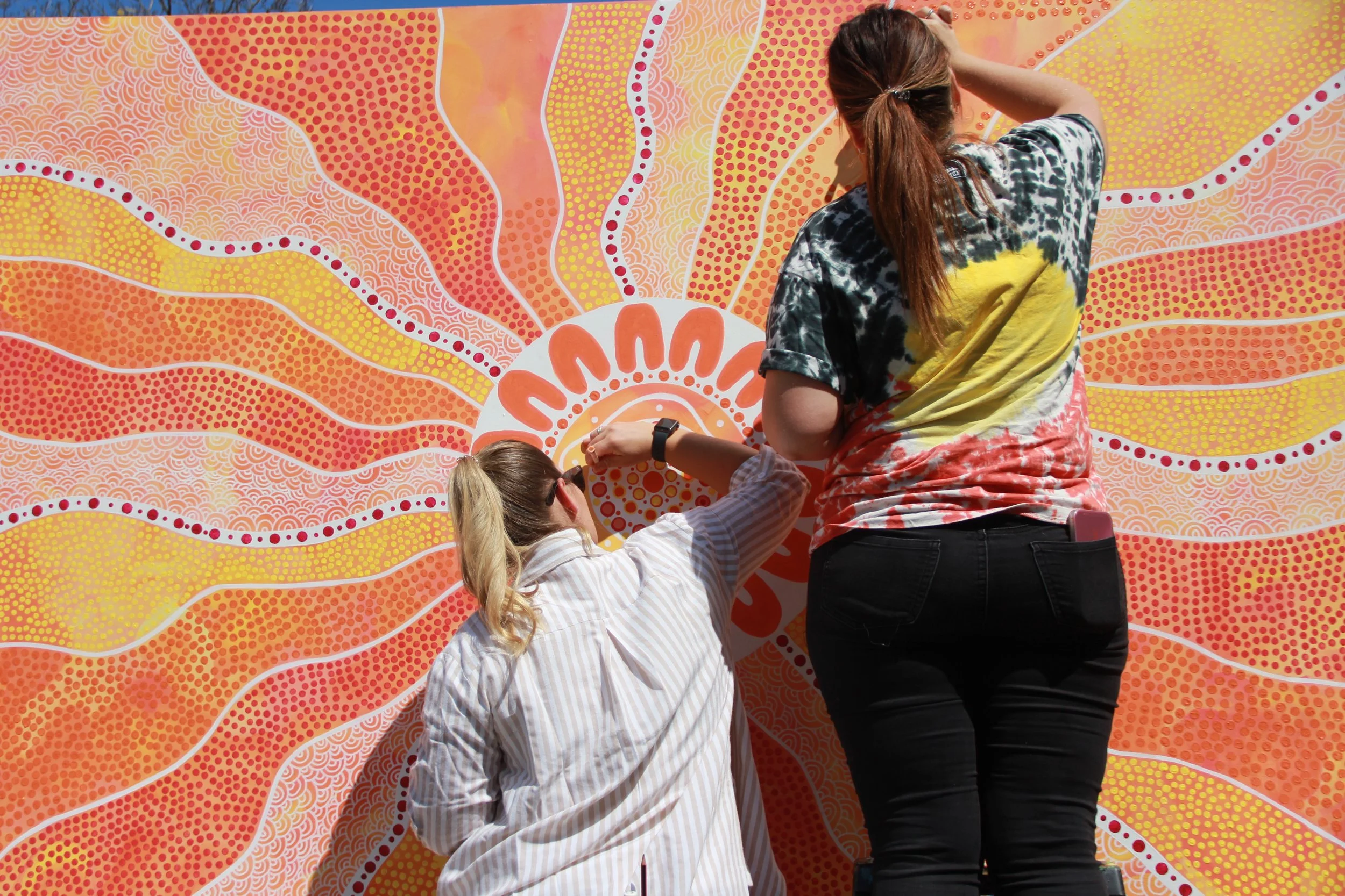 Two women working on a large, colorful mural featuring orange, yellow, and red patterns and a white circular design in the center.