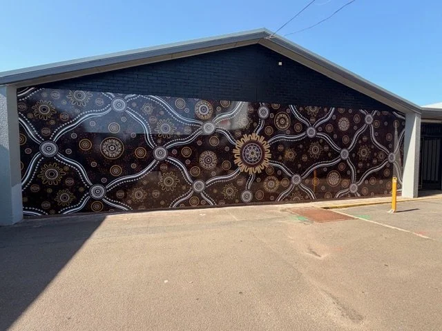 A mural with abstract patterns and circular designs in gold, white, and brown on a black background, painted on the side of a building.