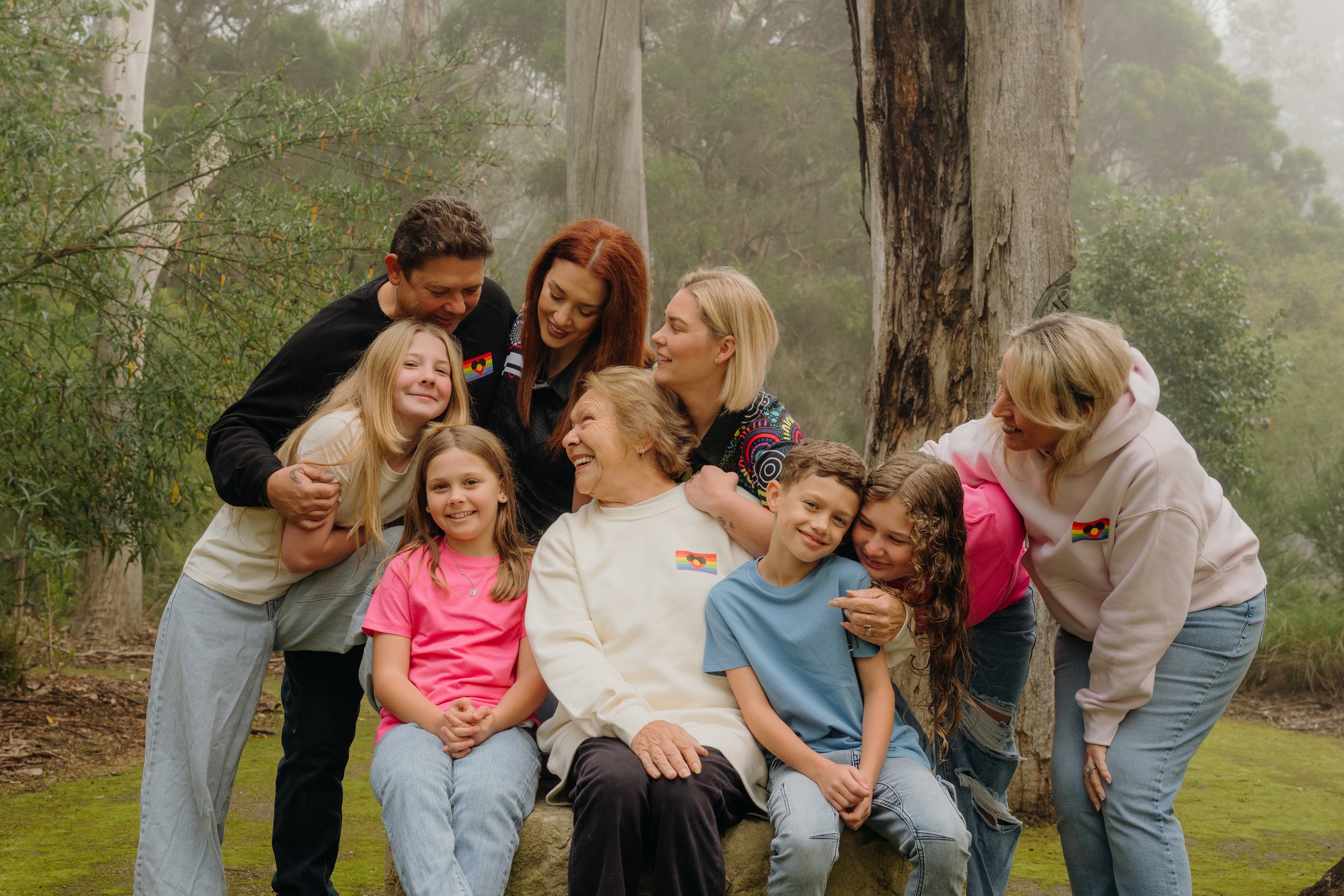 Family gathering of diverse generations smiling and hugging outdoors among trees, wearing rainbow-themed clothing.